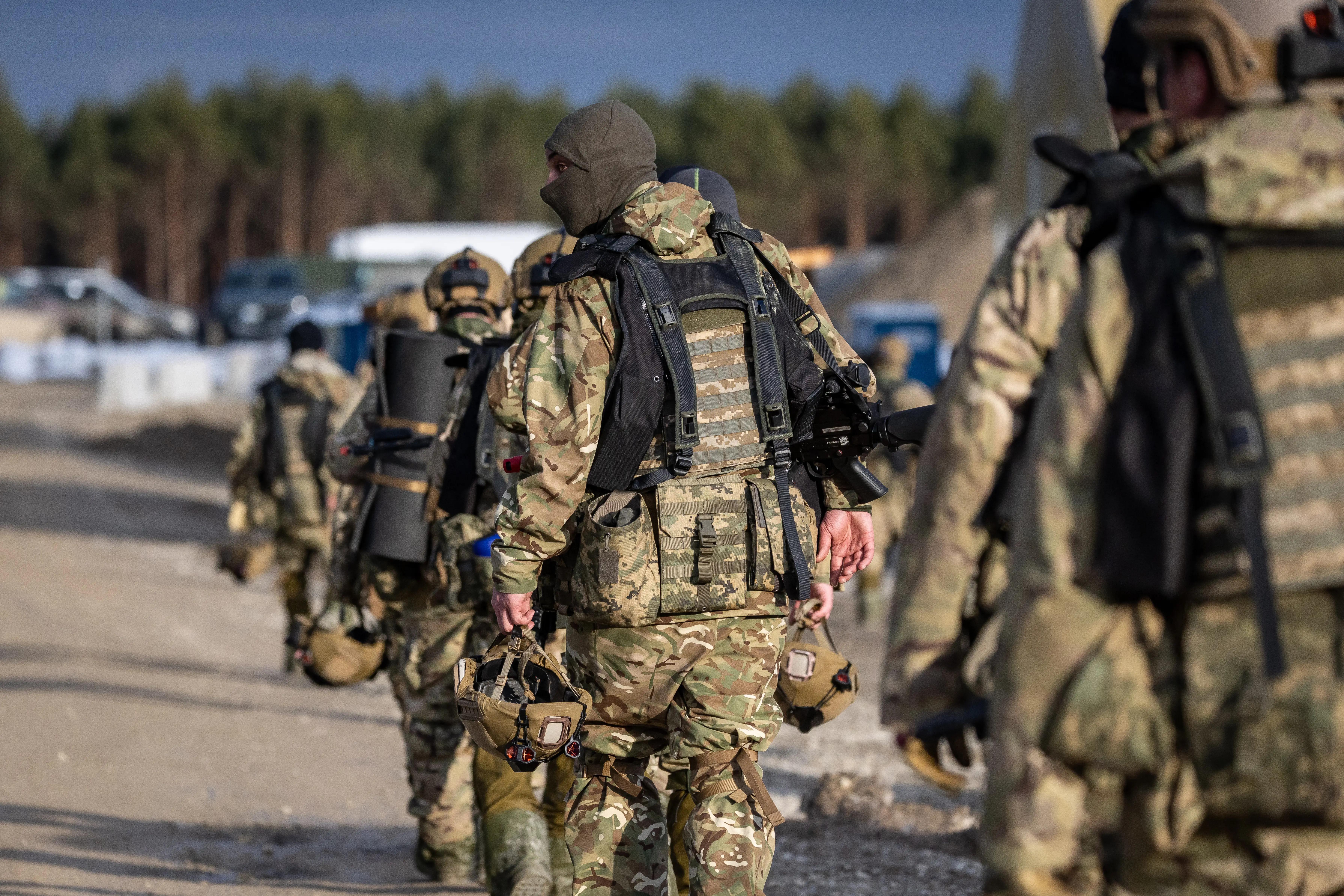 Camouflaged military personnel carrying gear walk along a paved road near a wooded area.