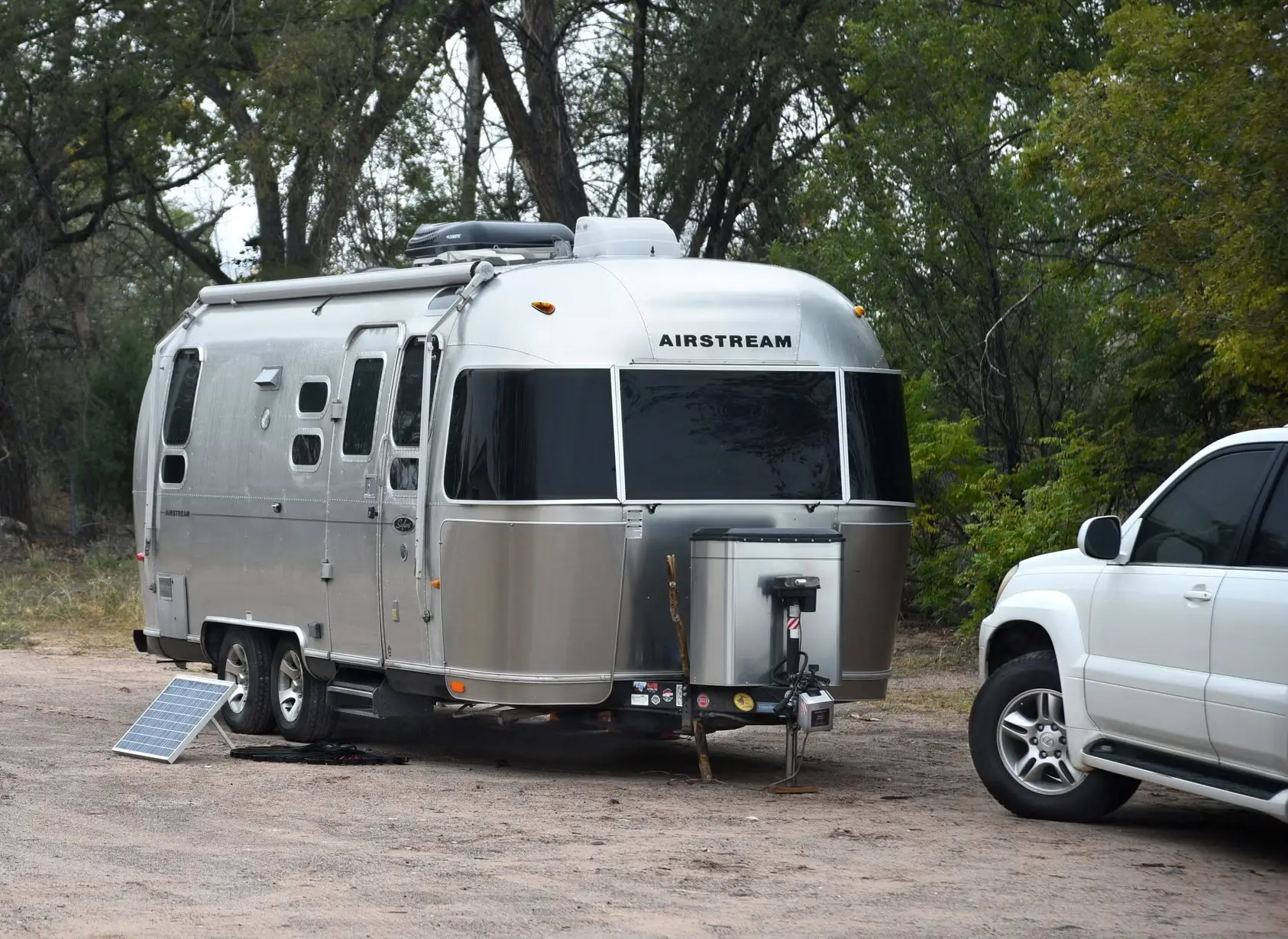 A silver Airstream travel trailer parked in a wooded campsite beside a white SUV and portable solar panel.