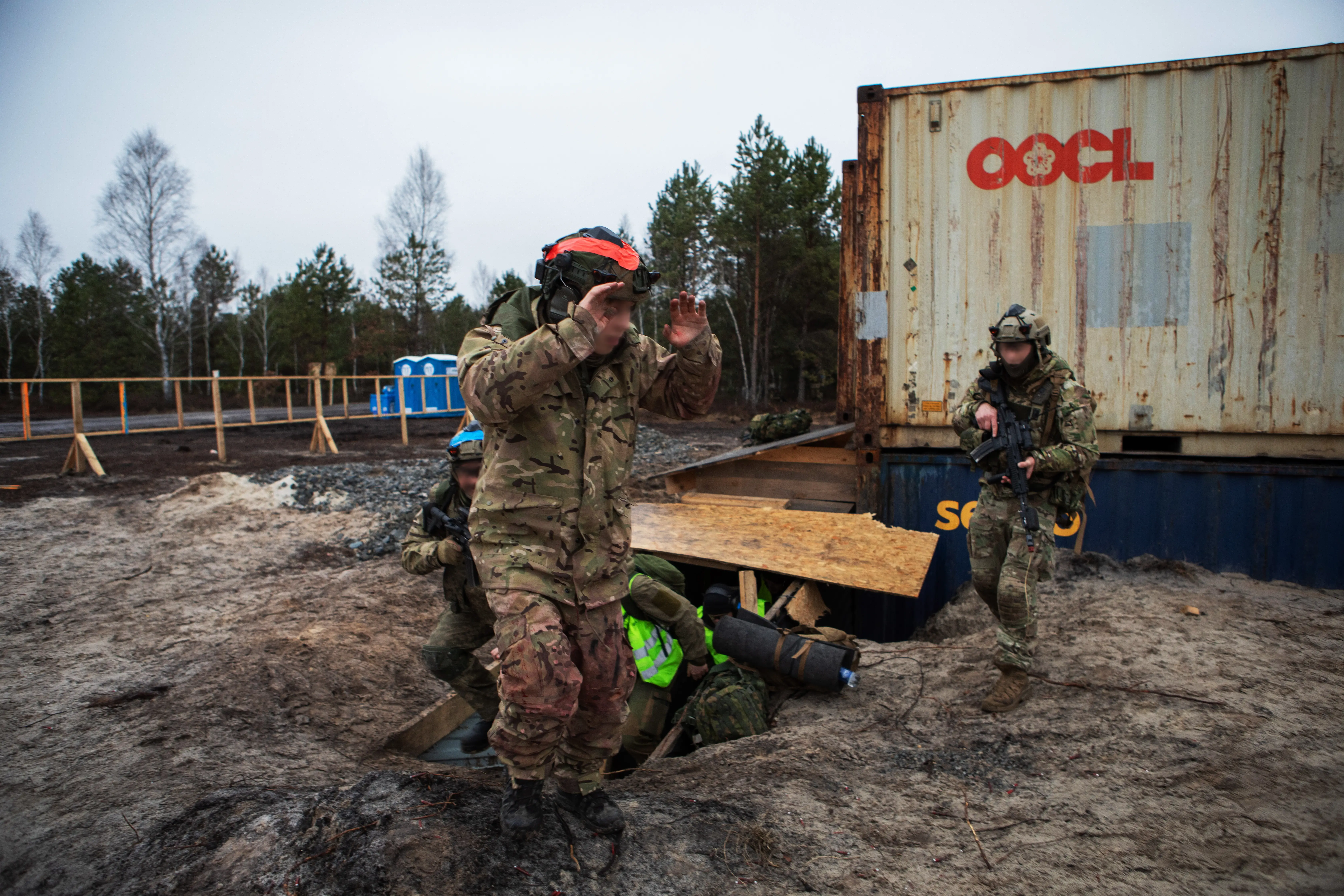 Camouflaged soldiers train around a dugout beside a rusted OOCL shipping container in a muddy wooded area.