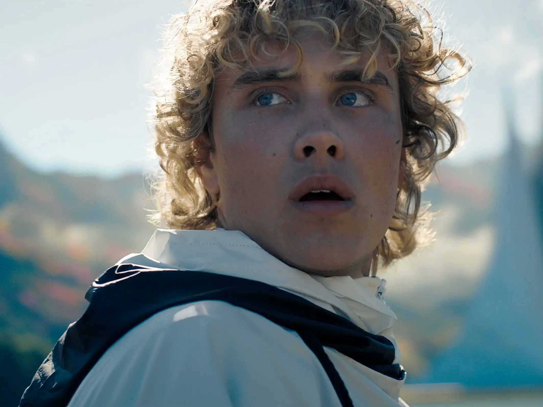 Curly-haired person in a white jacket looks upward near futuristic spires against a mountainous landscape.