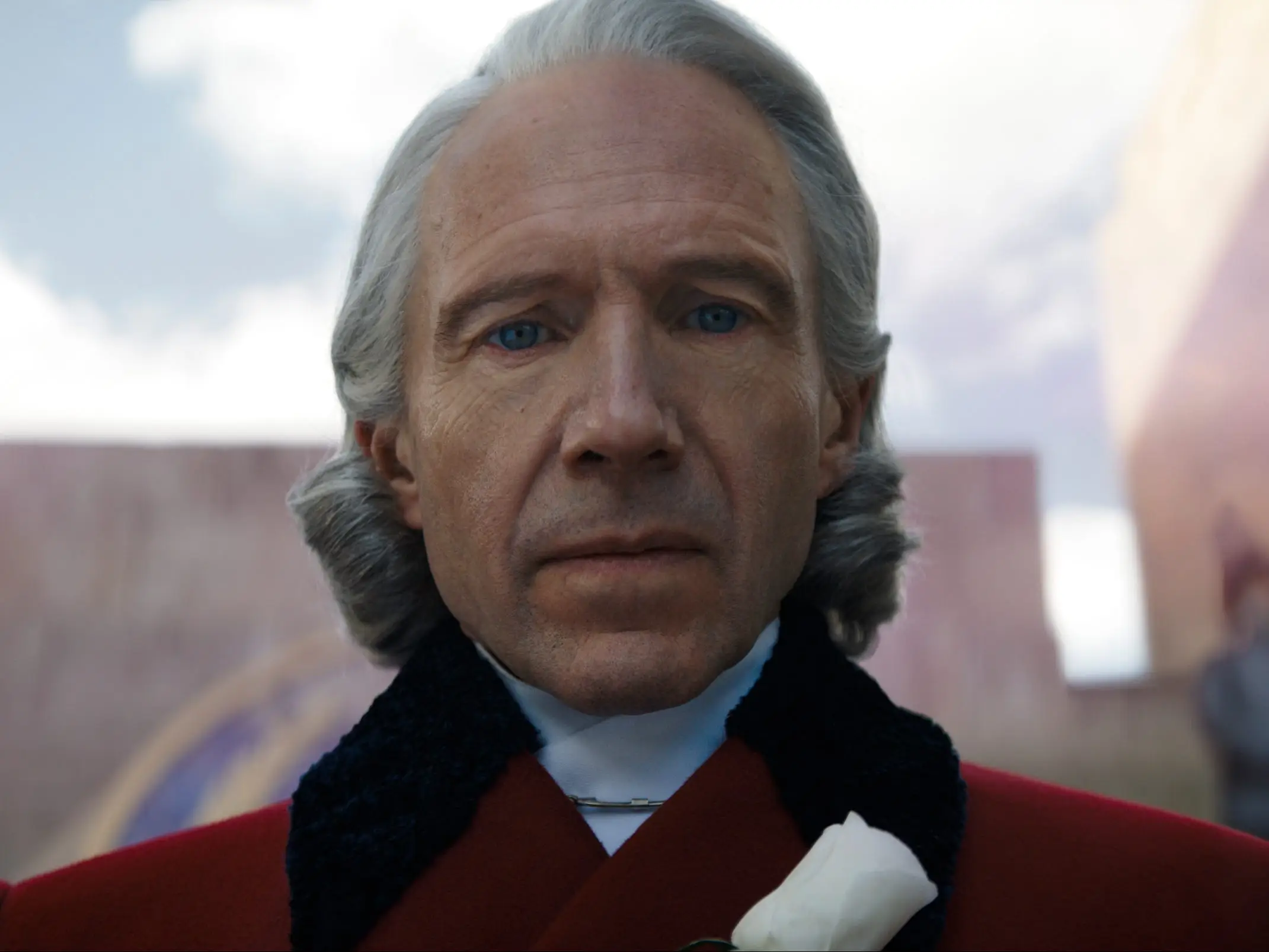 Ralph Fiennes as Coriolanus Snow stands in a red coat before blurred guards and monumental walls.