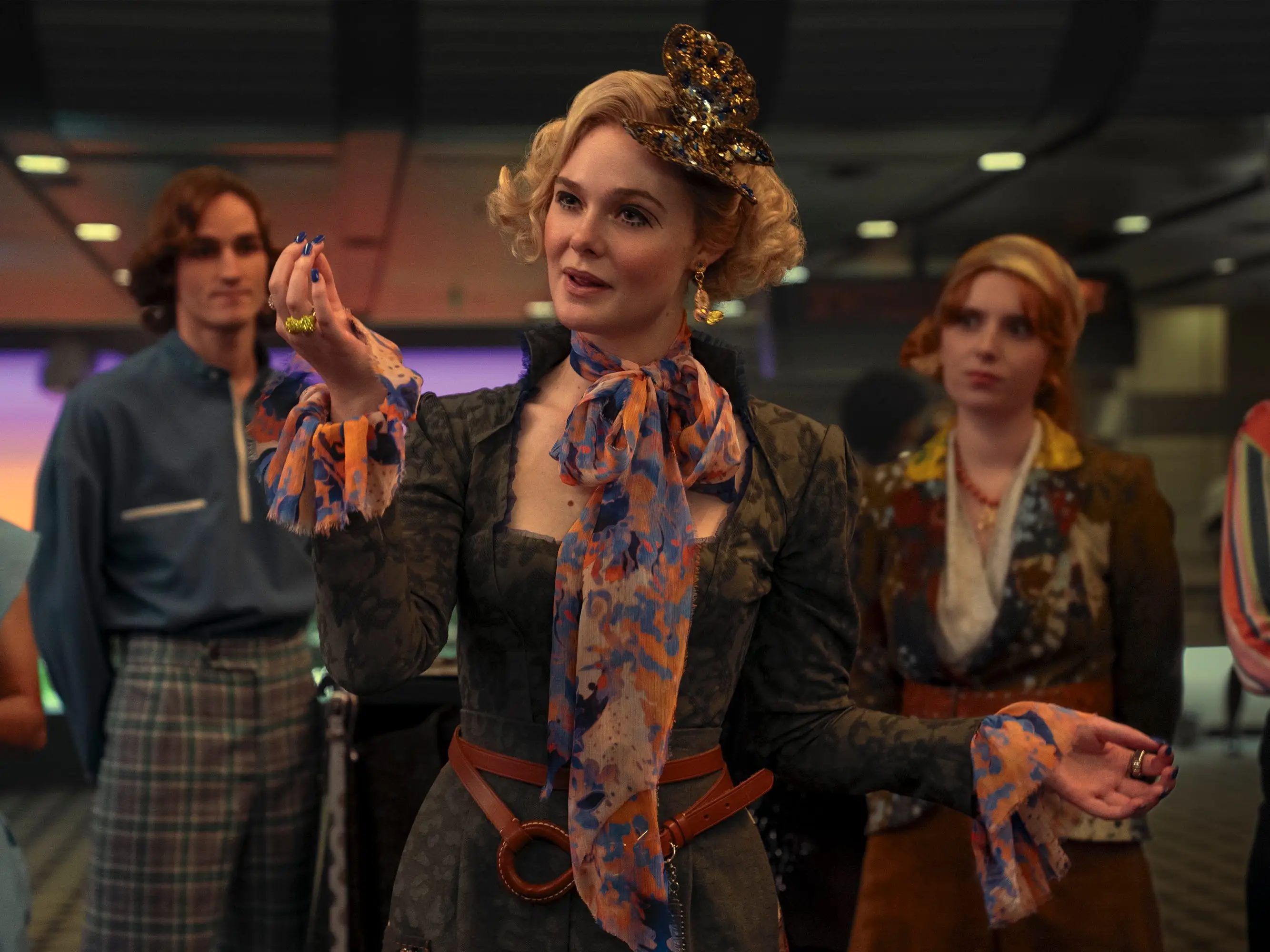Costumed group stands indoors as Elle Fanning as Effie Trinket with a decorative headpiece gestures toward the camera.