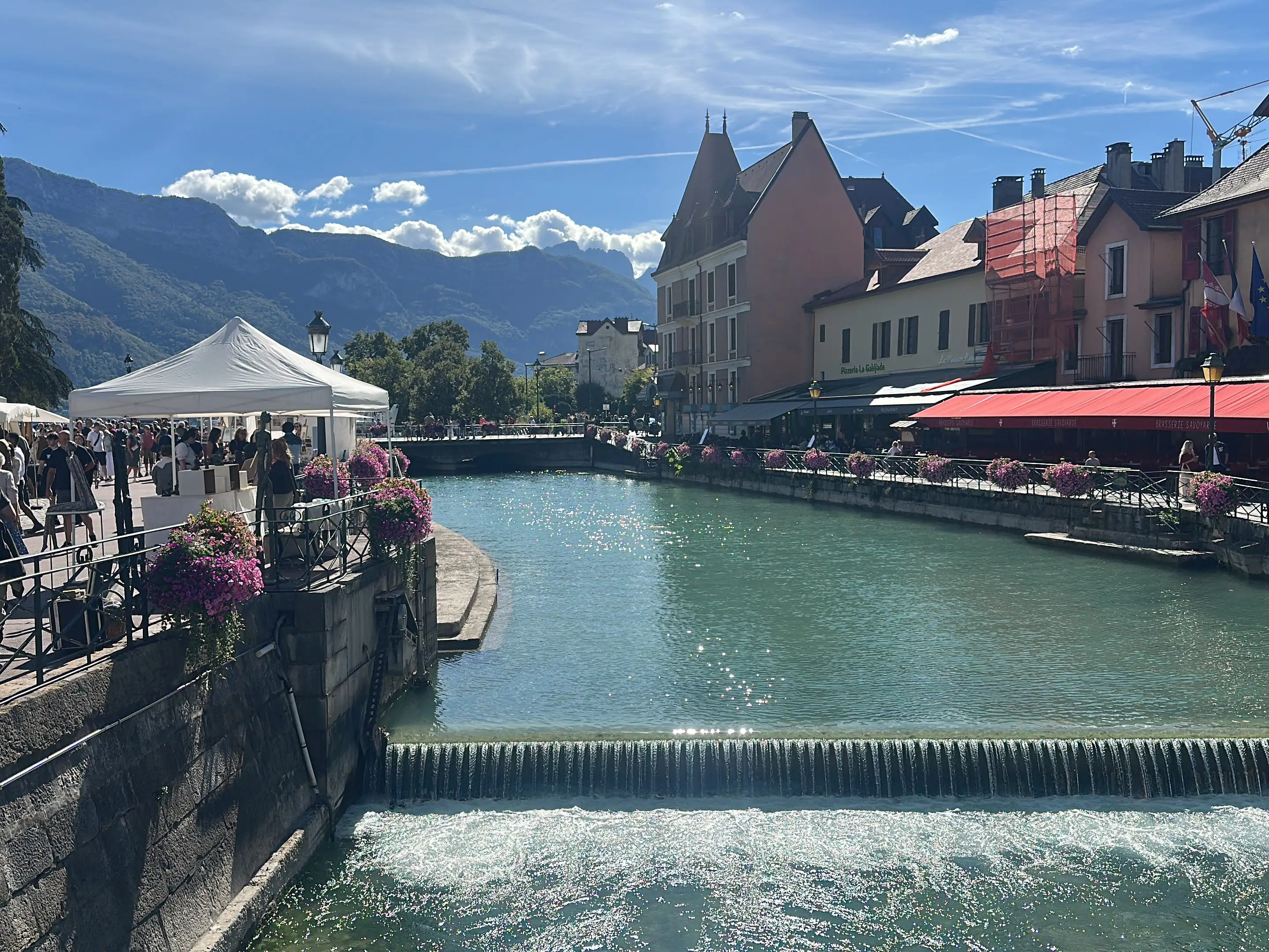 Canal-side pedestrian area with market tents, flower railings, cafés, flags, and alpine mountains under a sunny sky.