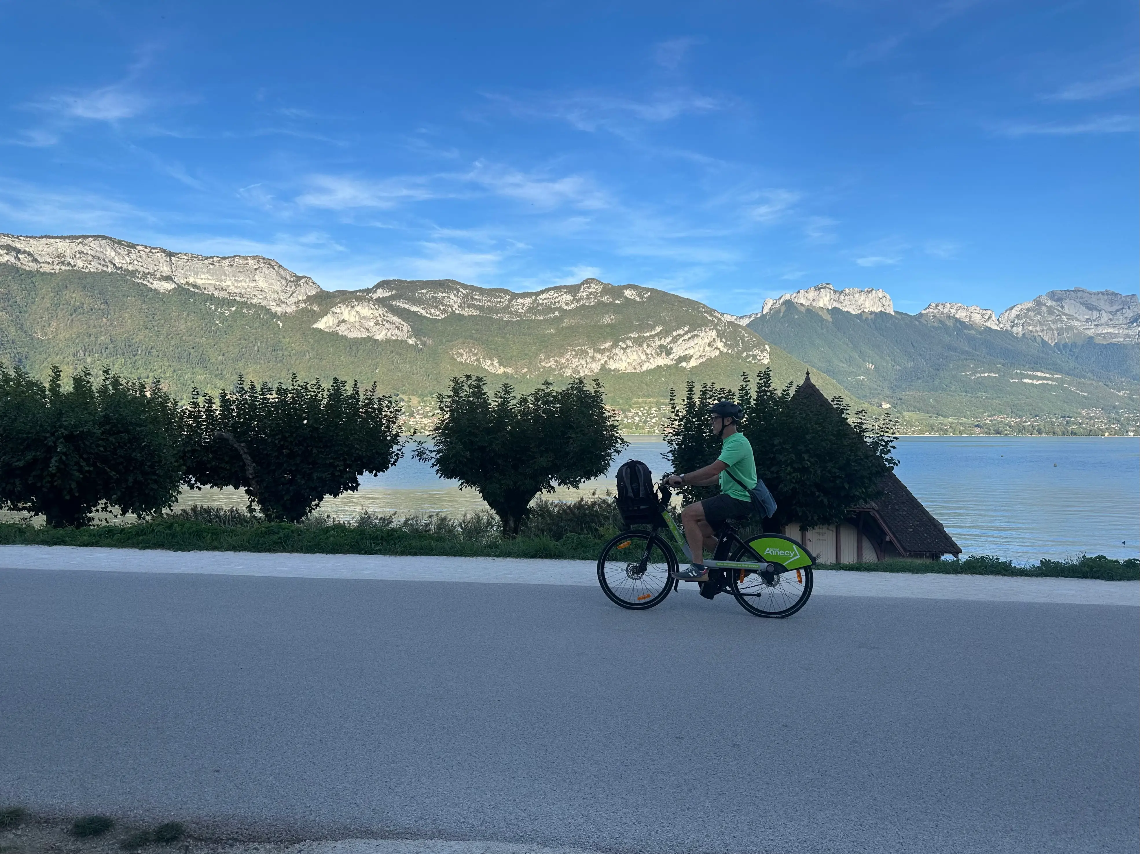 Cyclist rides a Vélonecy bicycle on a lakeside road with mountains and trees in the background.