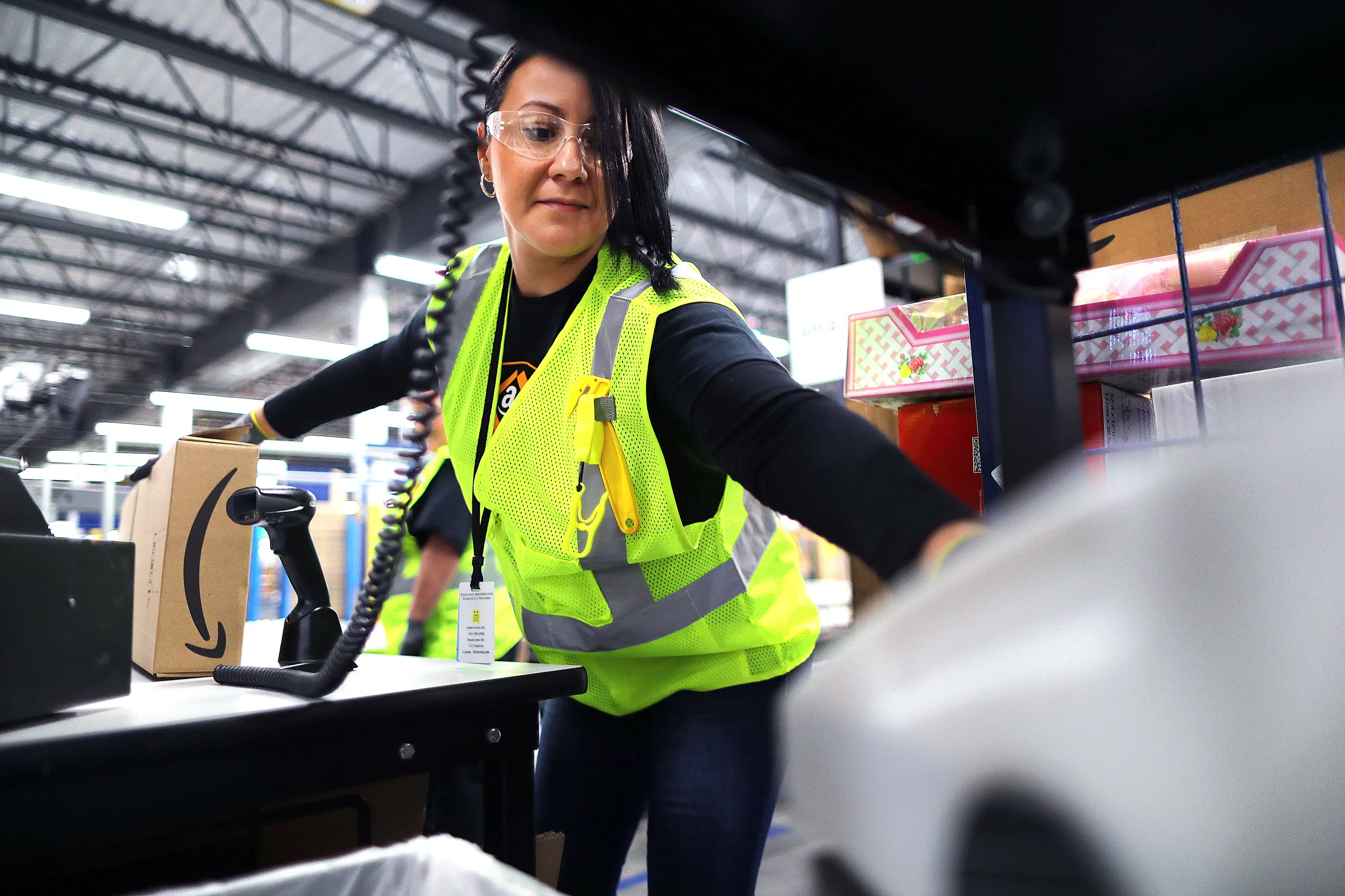 Worker in a high-visibility vest reaches across a workstation in a warehouse with an Amazon-branded box nearby.