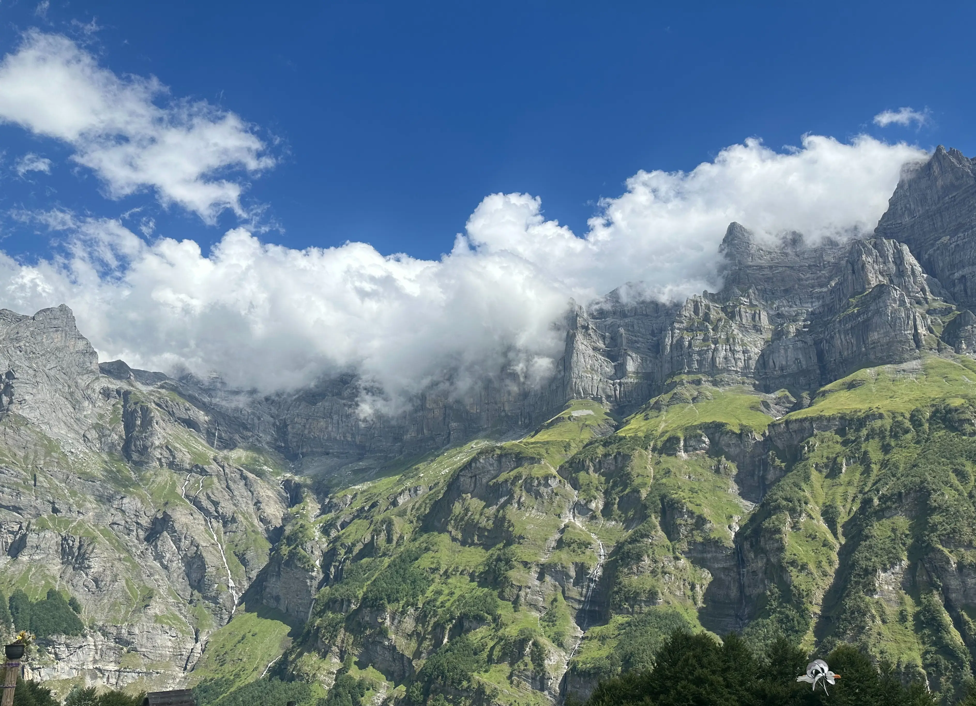 Clouds drift across rugged gray mountain cliffs above green slopes and small waterfalls under a blue sky.