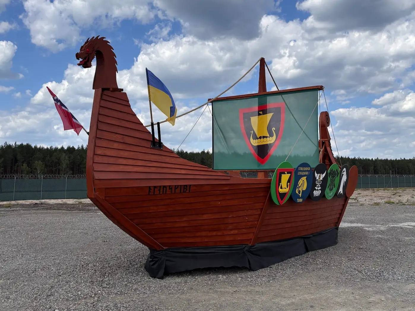 Wooden Viking-style boat display with flags and shields stands on a gravel lot under a cloudy sky.