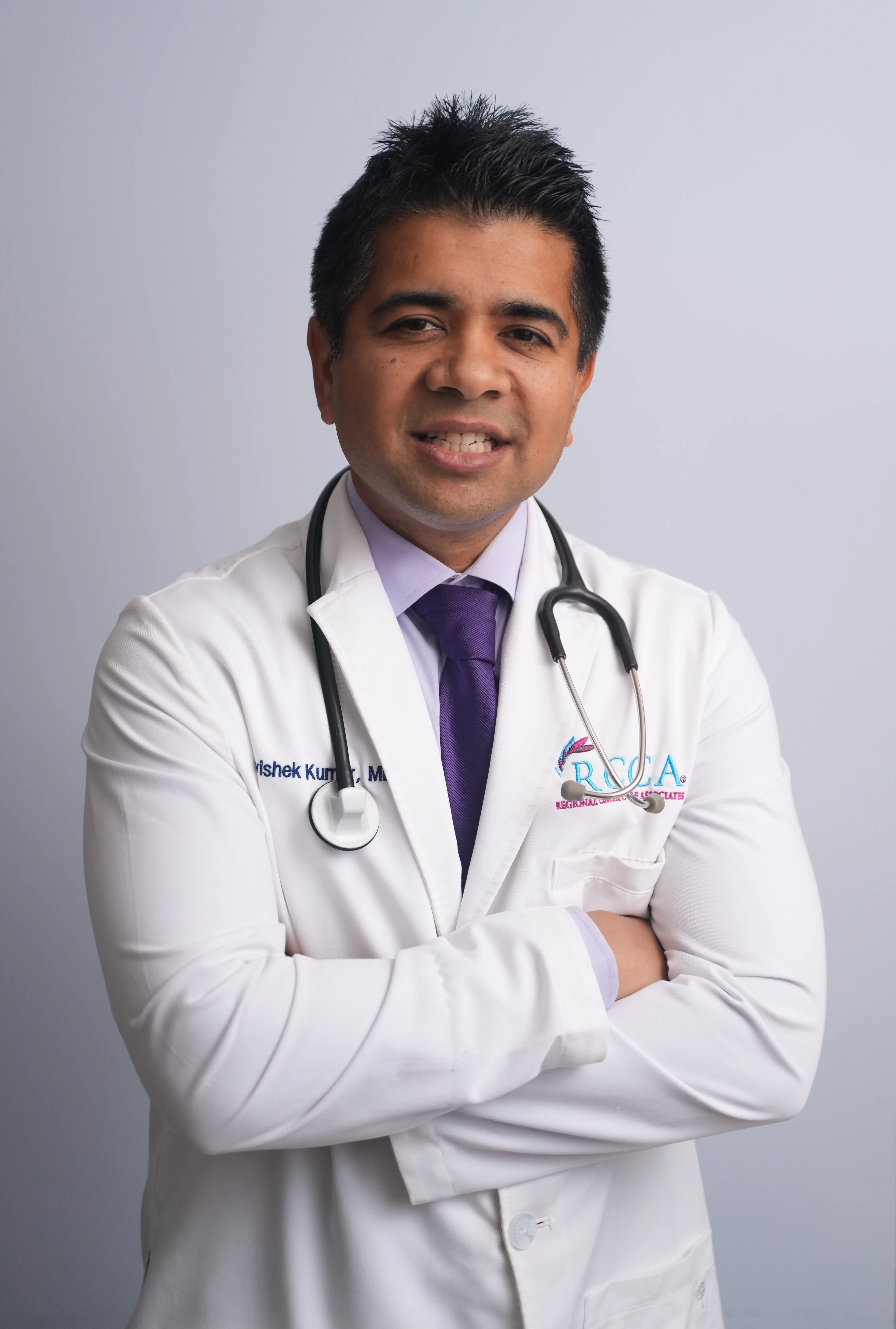 Medical professional in a white coat with stethoscope poses against a plain gray background.