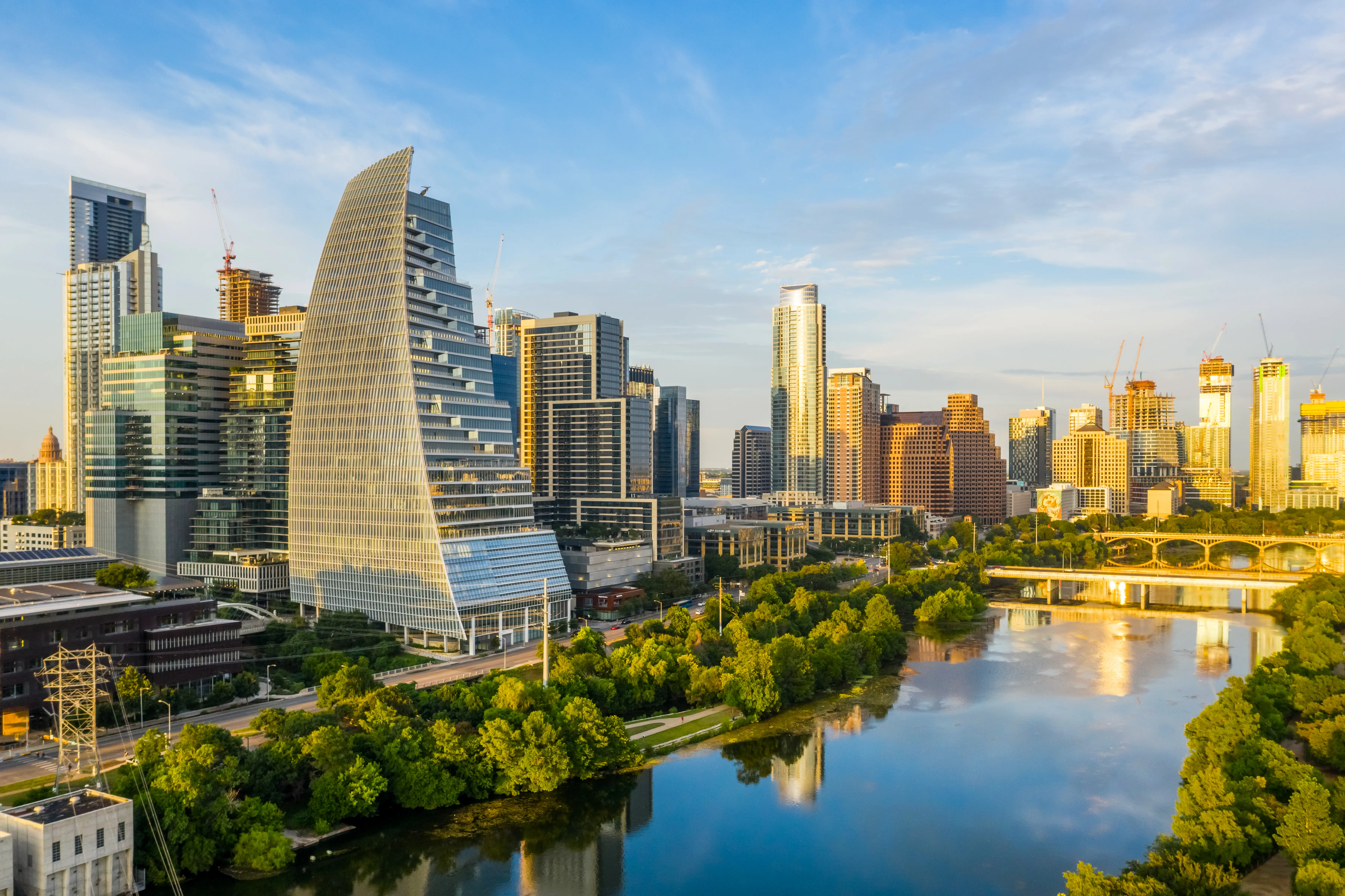 Modern high-rise skyline and a sail-shaped tower reflect beside a river at golden hour in Austin.