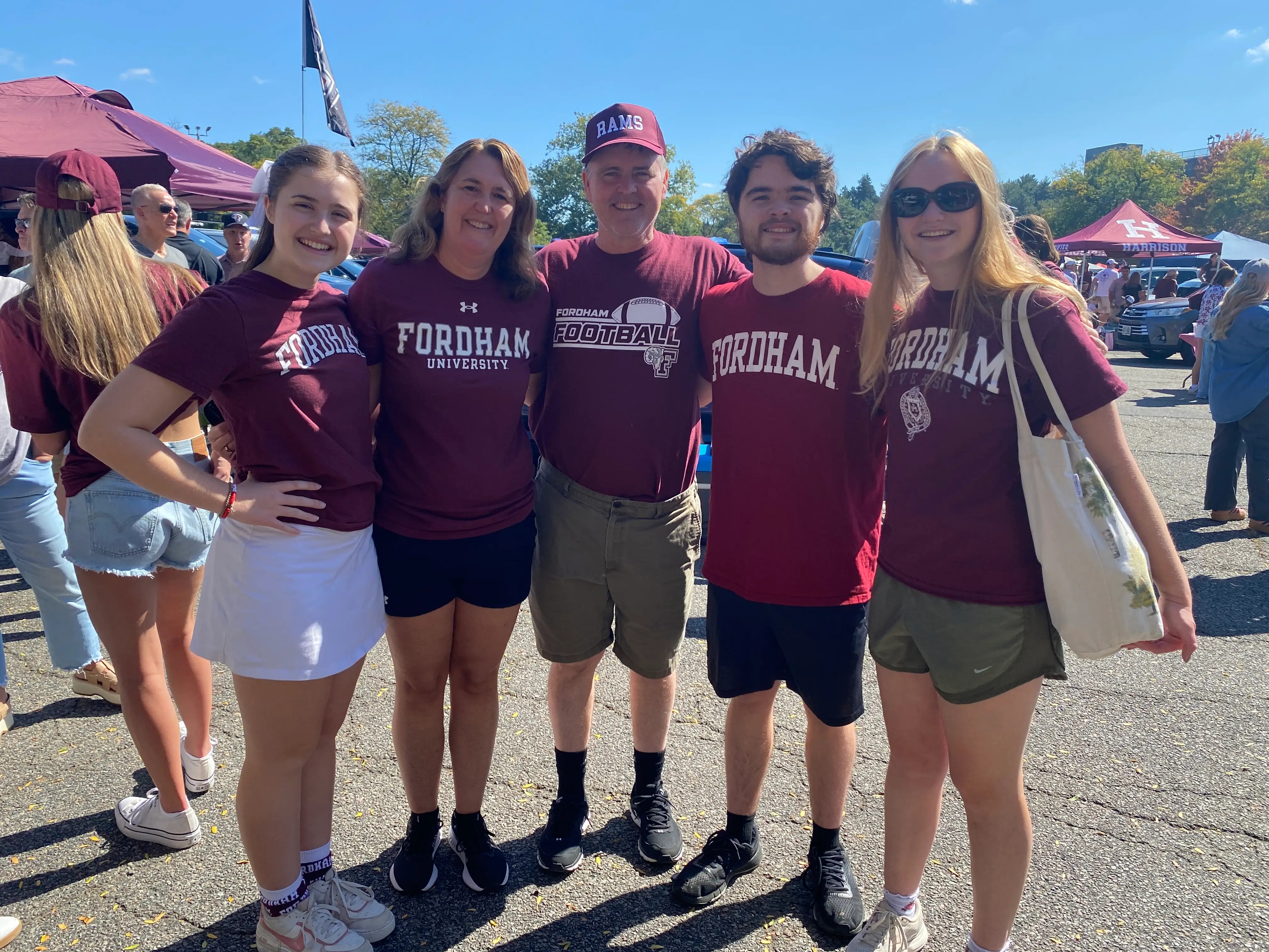 Five people in Fordham shirts pose together at a sunny outdoor tailgate with maroon tents.
