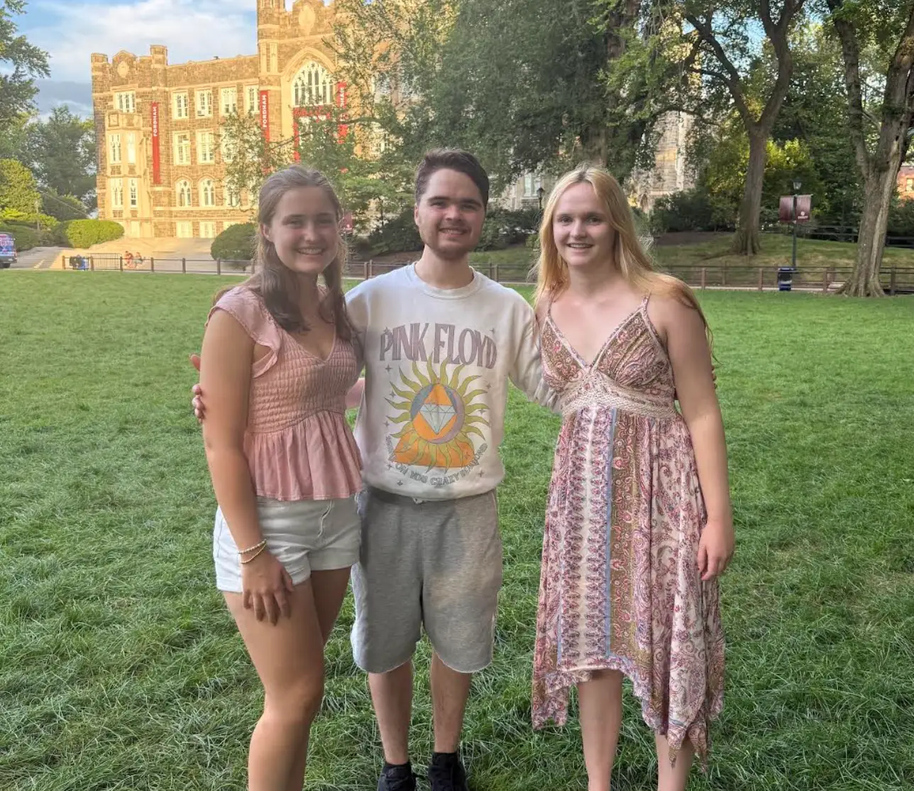 Three people pose on a grassy campus lawn in front of a Gothic-style building and trees.