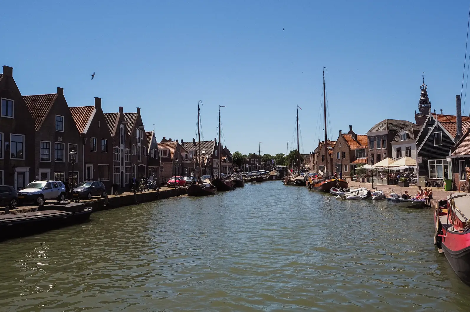 Canal lined with moored boats, brick waterfront buildings, parked cars, and café umbrellas under a clear blue sky.
