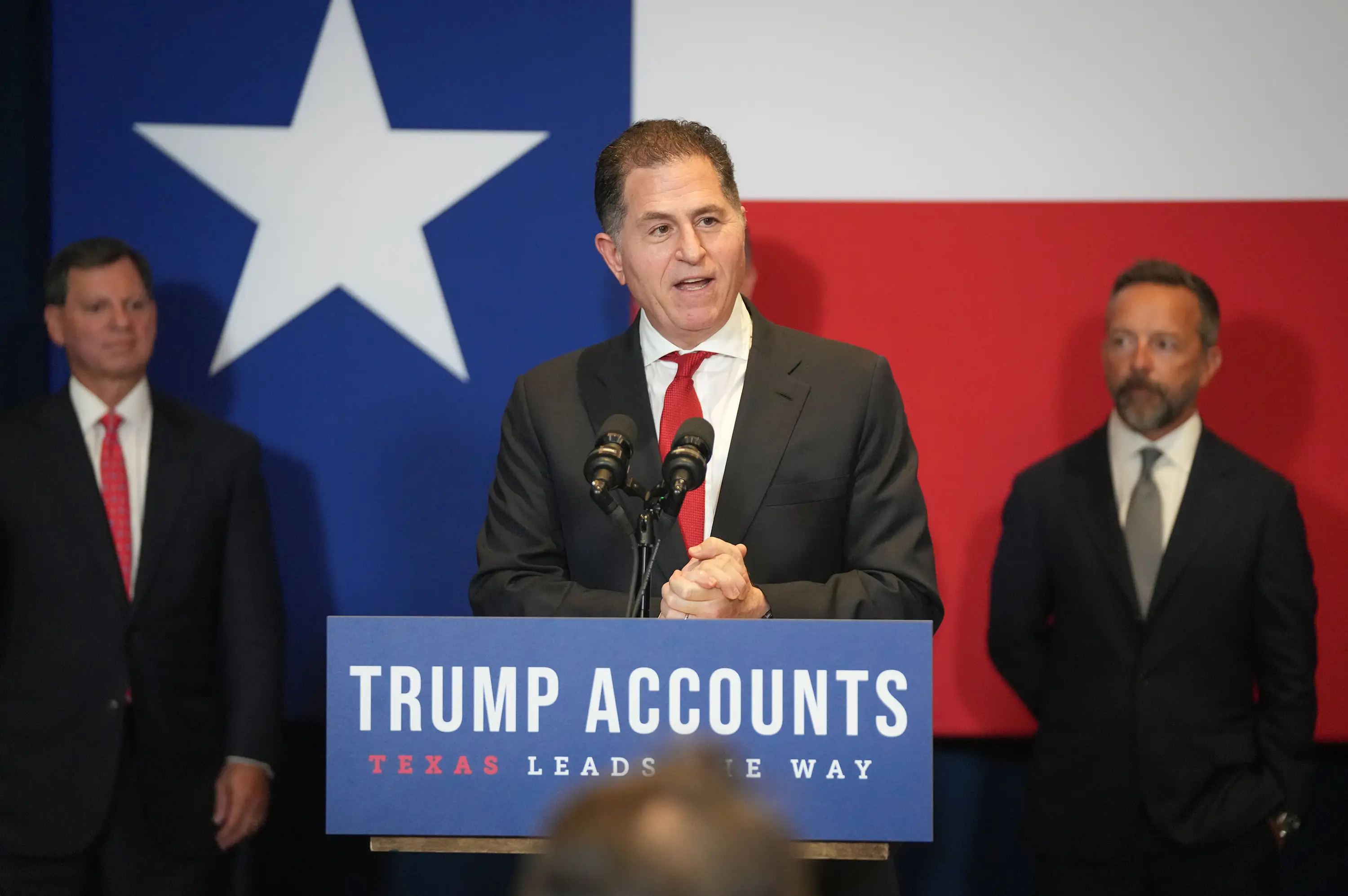 A man in a tuxedo speaks before a crowd, with a Texas flag in the background.