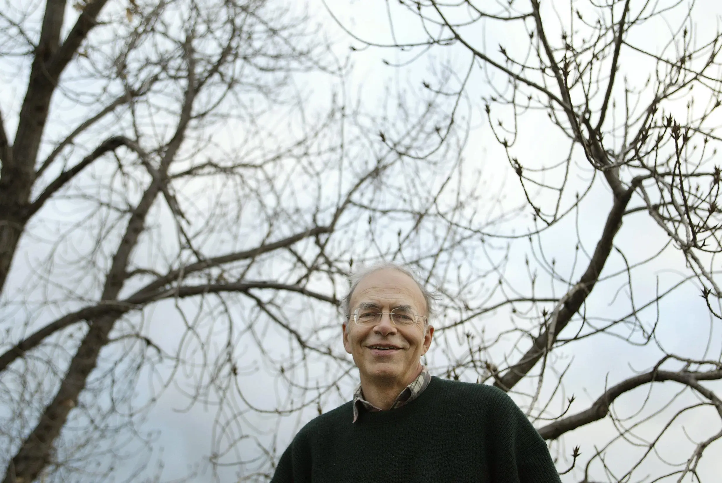 Smiling person wearing glasses stands outdoors beneath leafless tree branches against an overcast sky.