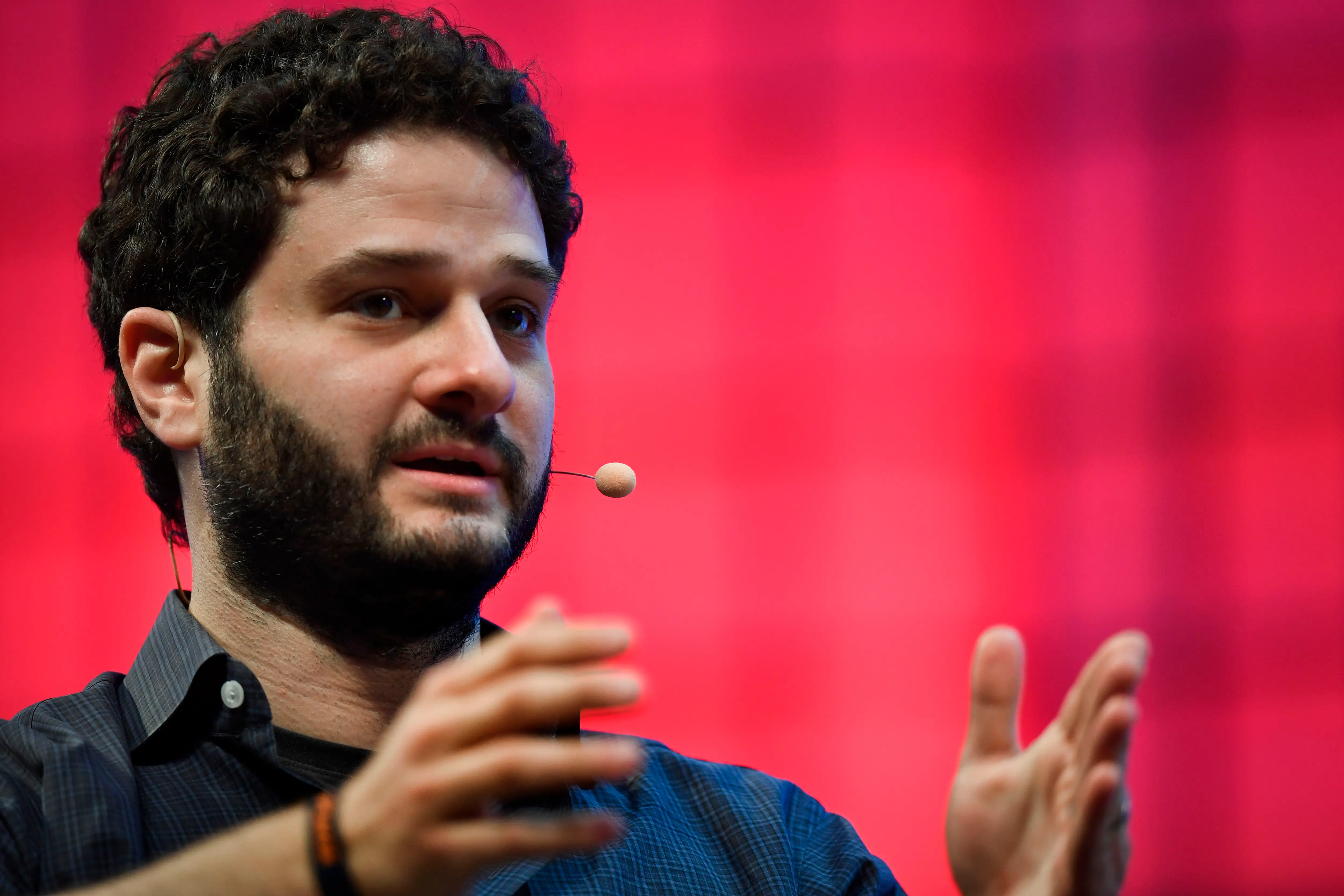 Speaker wearing a headset microphone gestures onstage against a red conference backdrop.