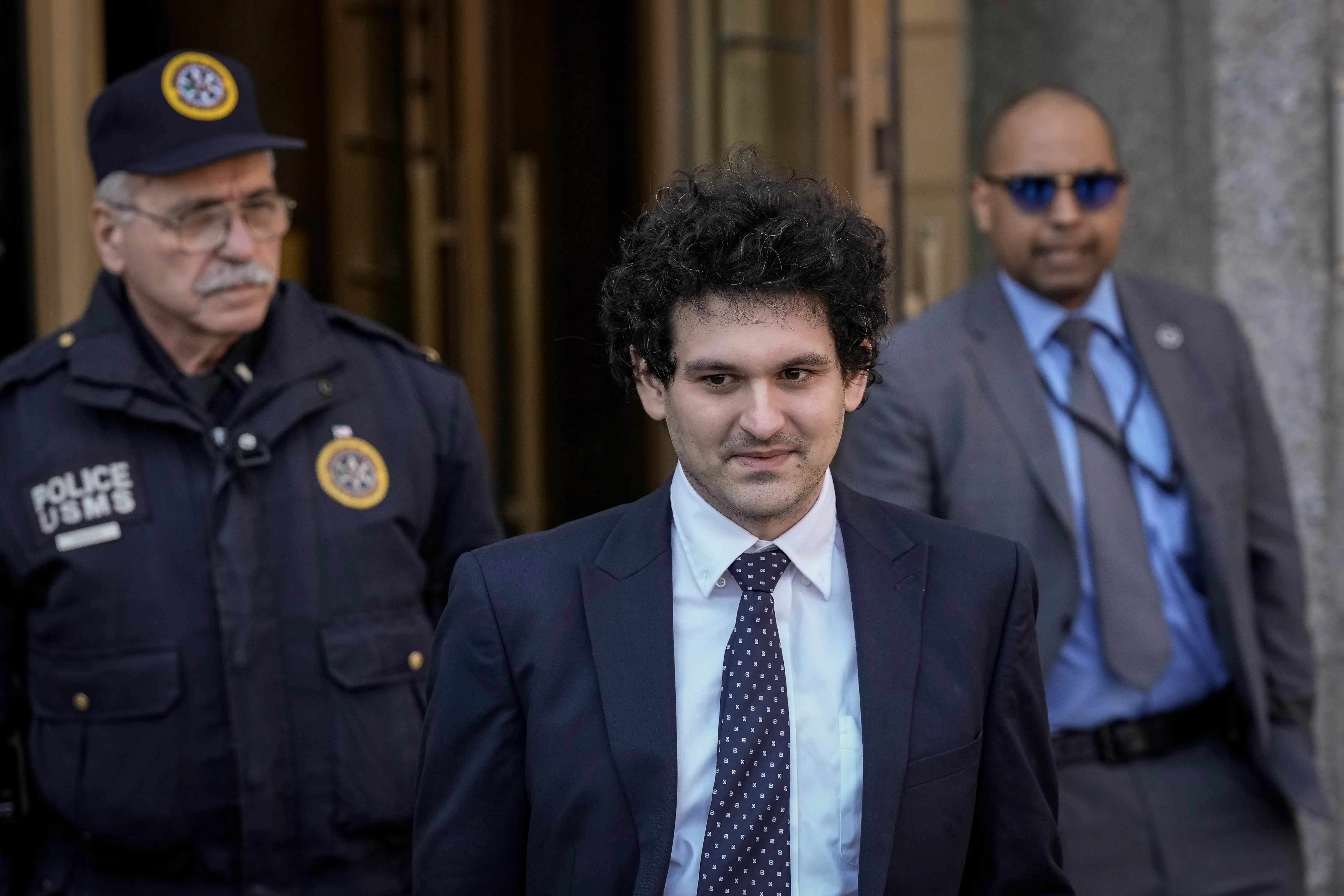 Sam Bankman-Fried walks outside a building flanked by uniformed court police and a suited security officer.
