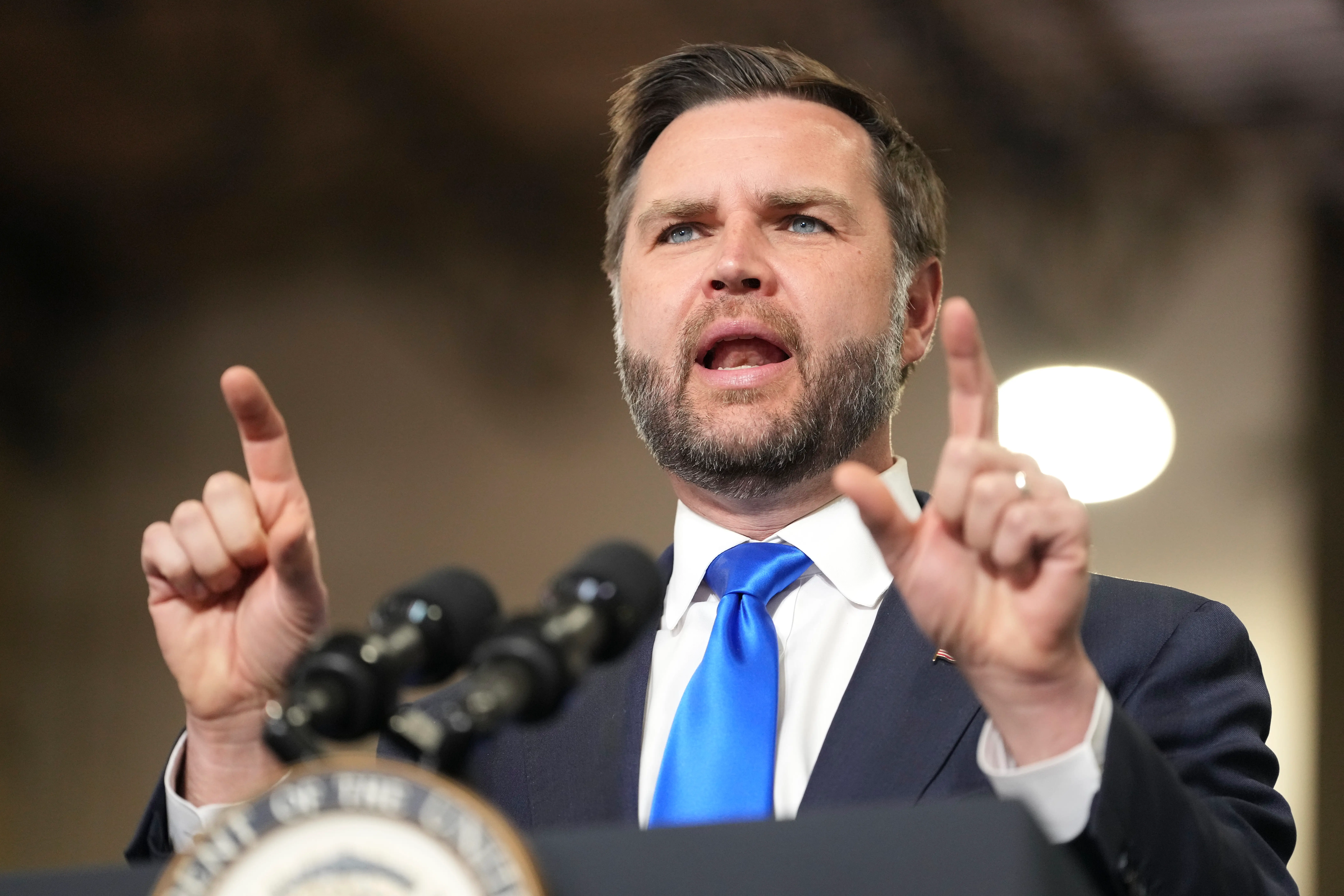 J.D. Vance speaks at a podium with microphones, raising both index fingers during an indoor public address.