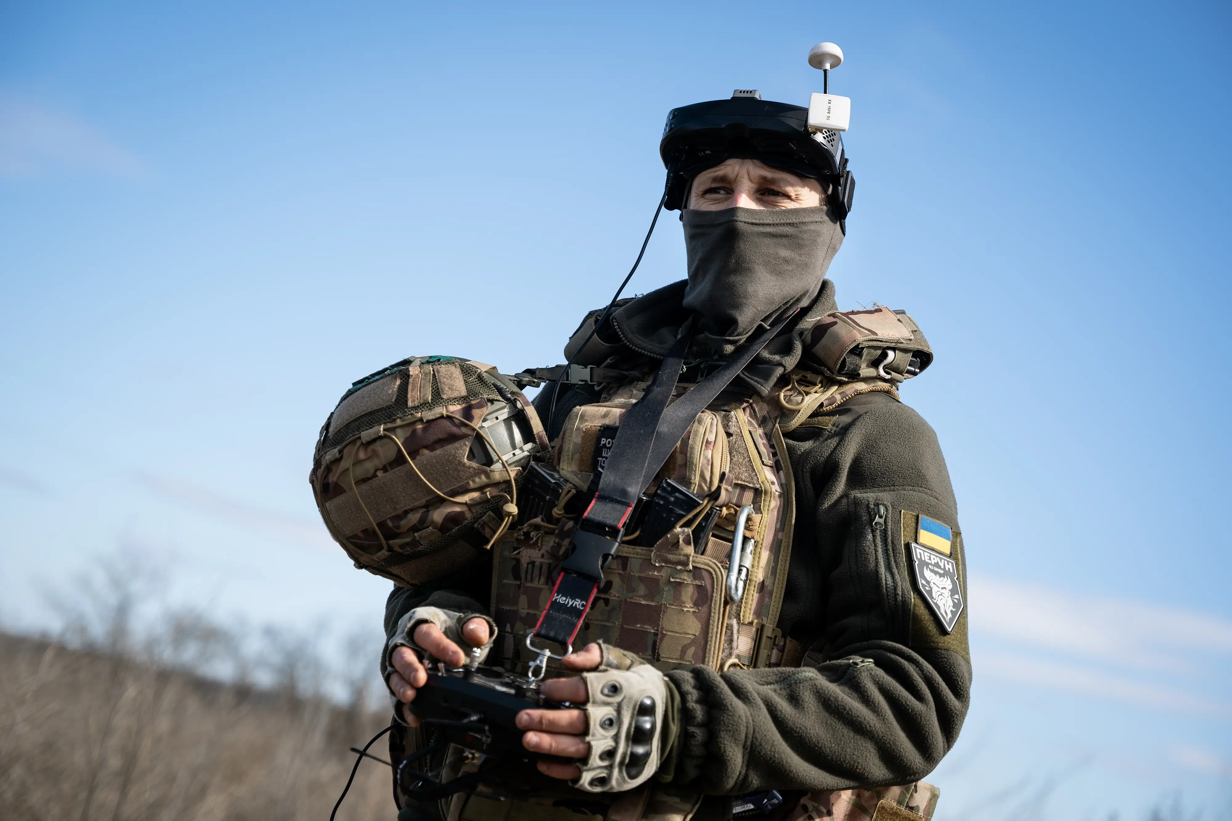Masked soldier in tactical gear holds a helmet and drone controller beneath a clear blue sky.