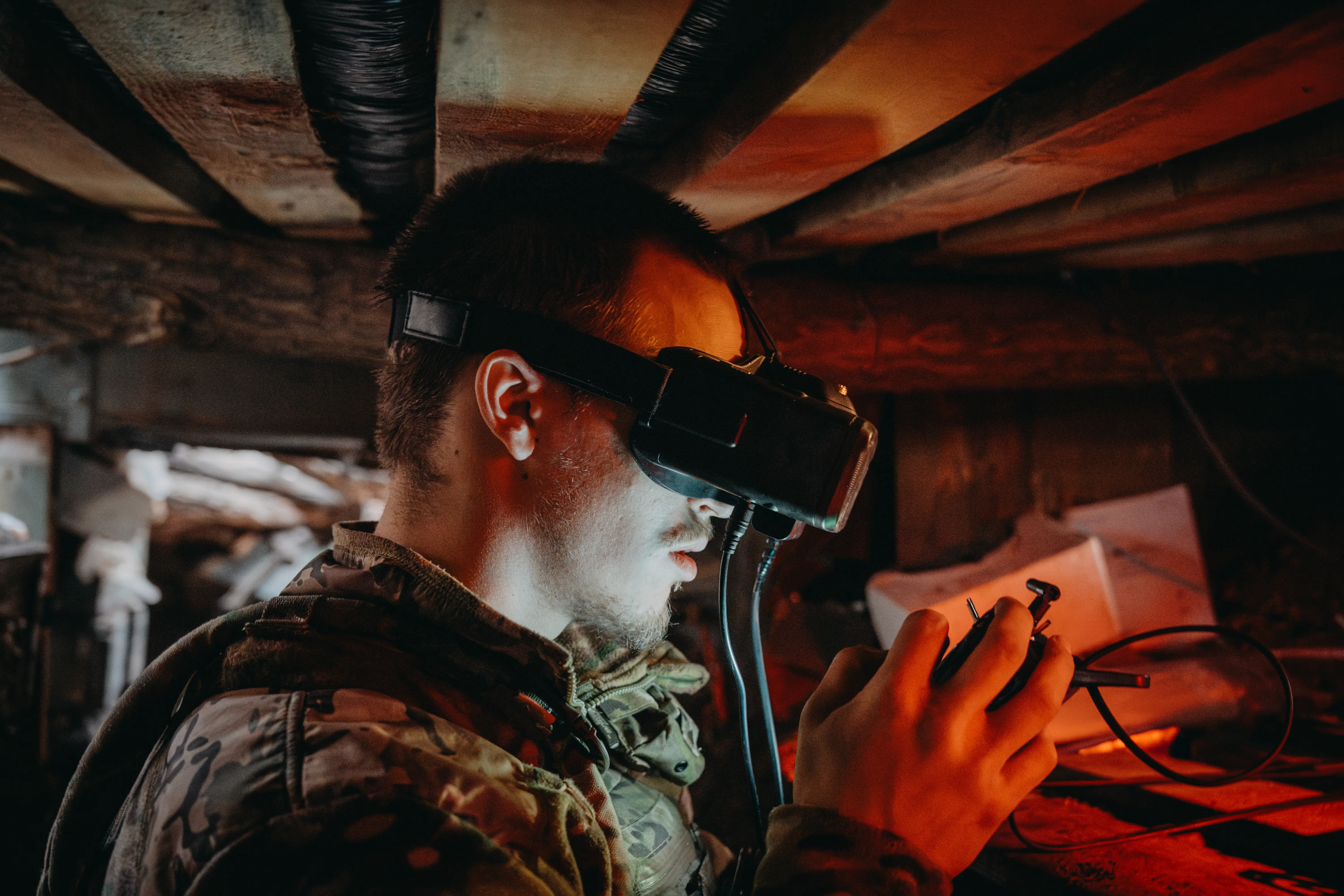 Camouflage-clad person uses a virtual reality headset and handheld controller in a dim, confined workspace.