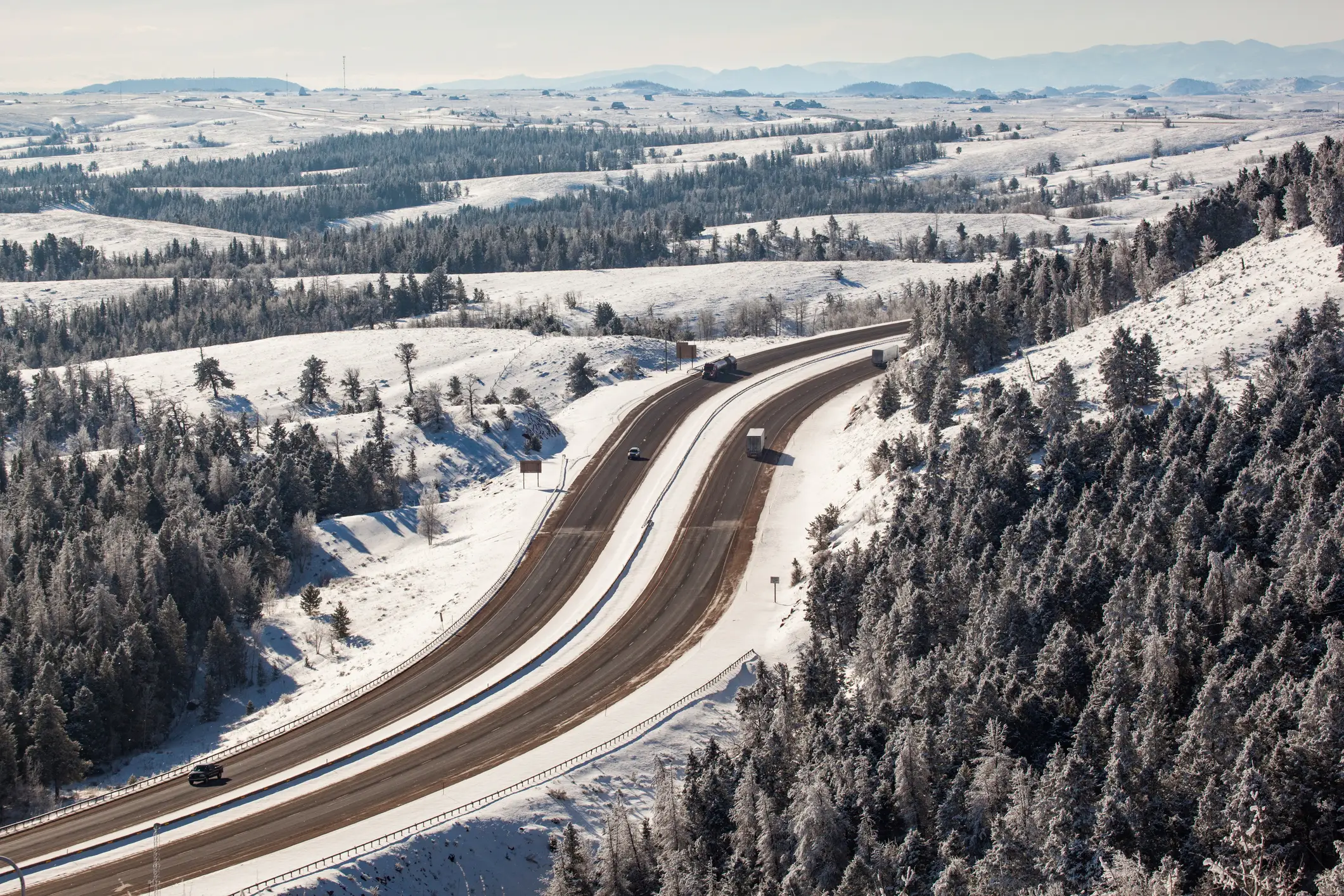 Snow-covered rural highway curves through forested hills with several vehicles under a pale winter sky.