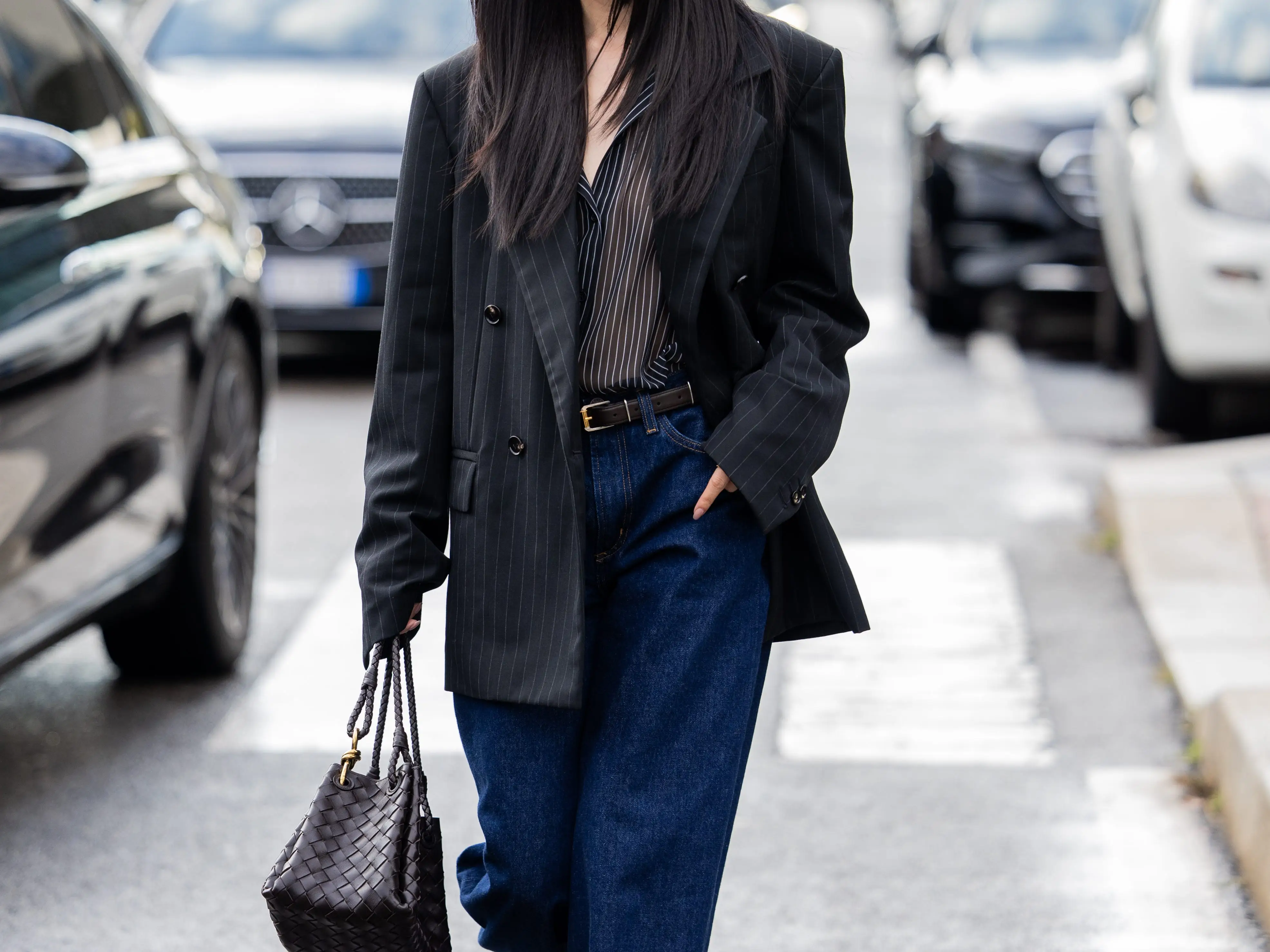 A woman in a dark blazer and jeans walks on a city street carrying a woven black handbag.