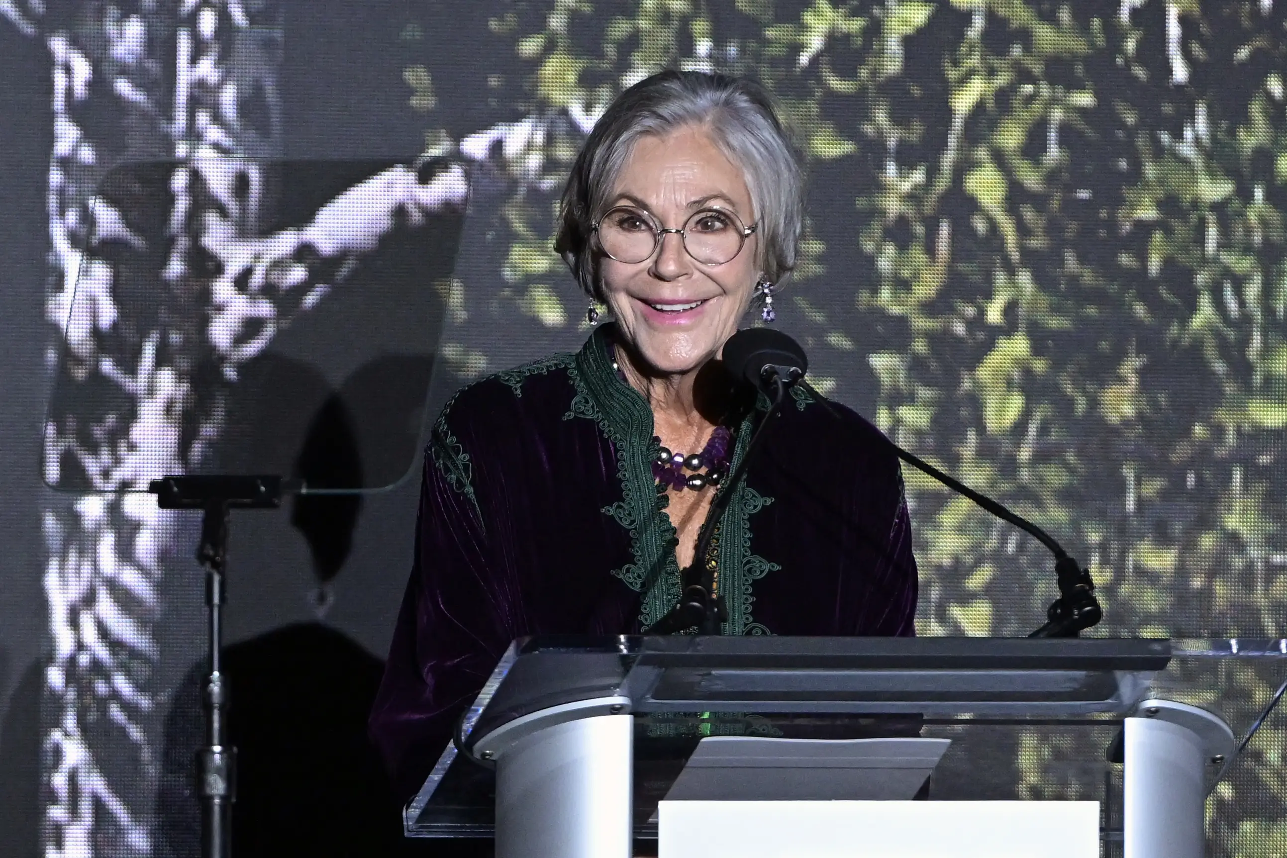 Alice Walton smiles while speaking at a clear podium on a stage with a leafy projected backdrop.