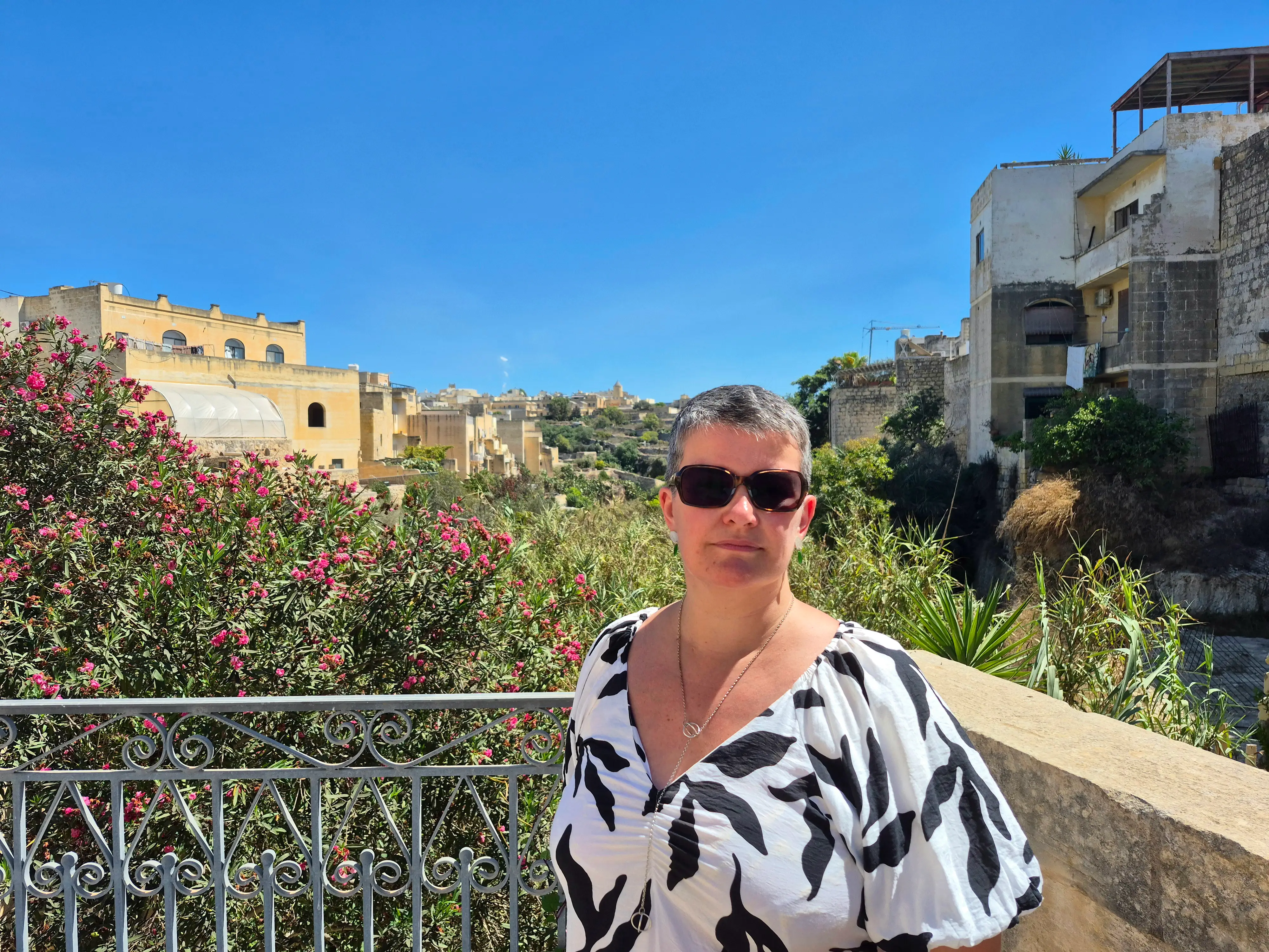 The writer standing on a balcony in Gozo, near buildings and pink flowers.