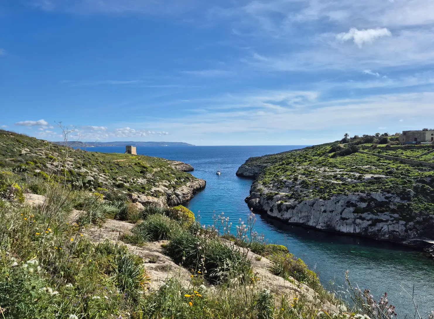 Rocky coastal inlet with turquoise water, green hillsides, a small boat, and blue sky in Gozo.