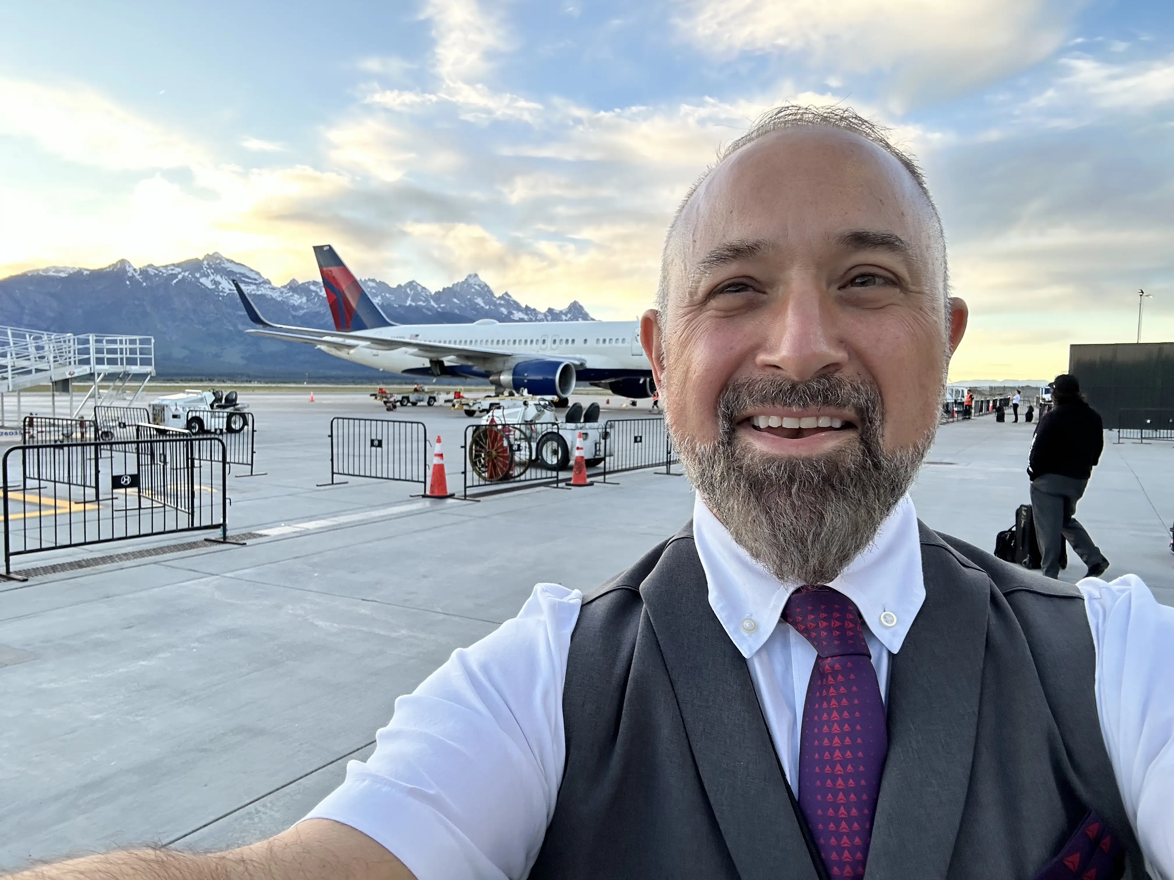 A flight attendant taking a selfie with a plane in the background.