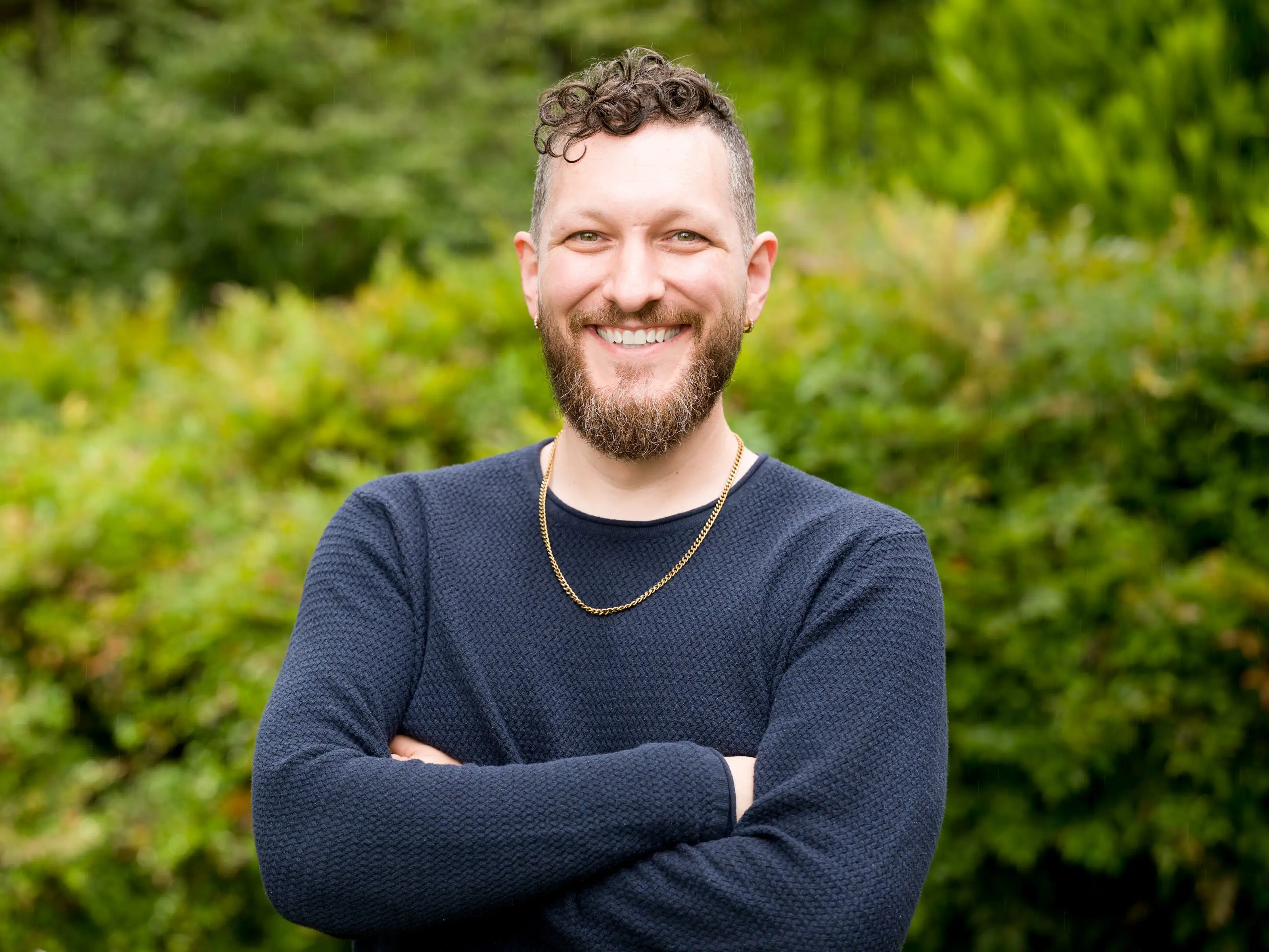 Smiling bearded person in a dark sweater stands with arms crossed in front of green foliage.