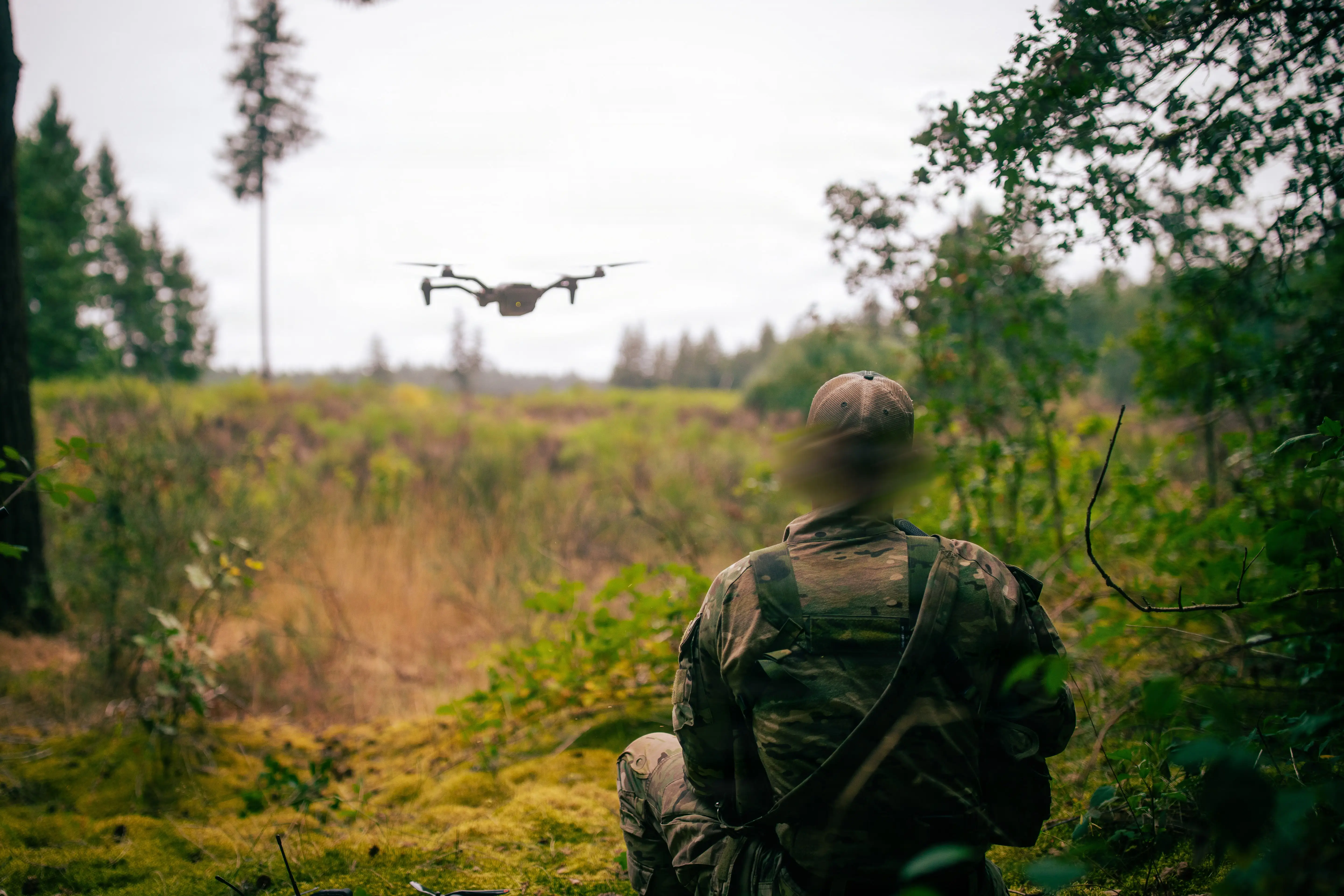 A US Army soldier sits in a grassy field flying a drone against an overcast sky.