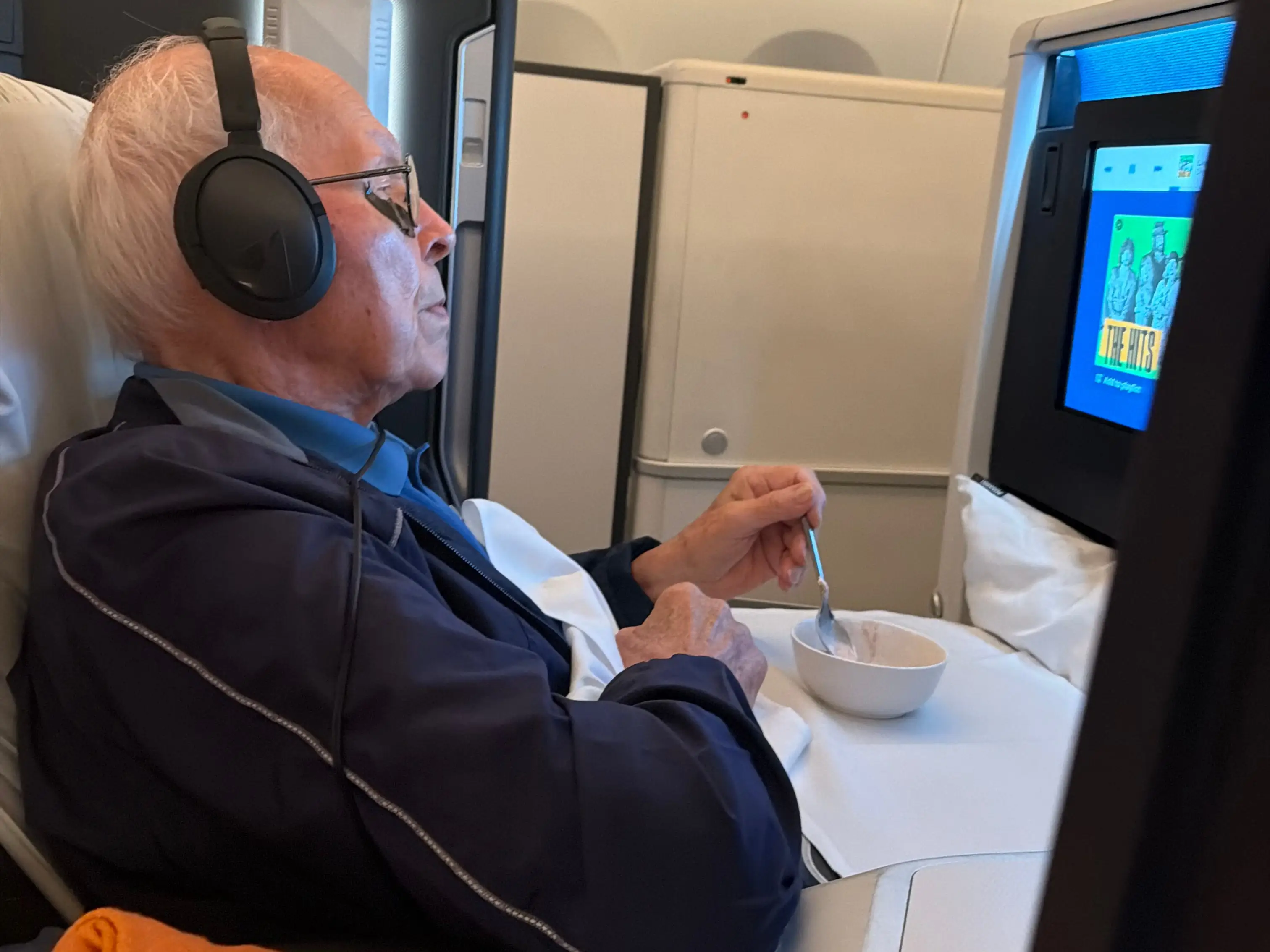 An older man in an airplane seat is eating an ice cream sundae while watching the seat-back TV.