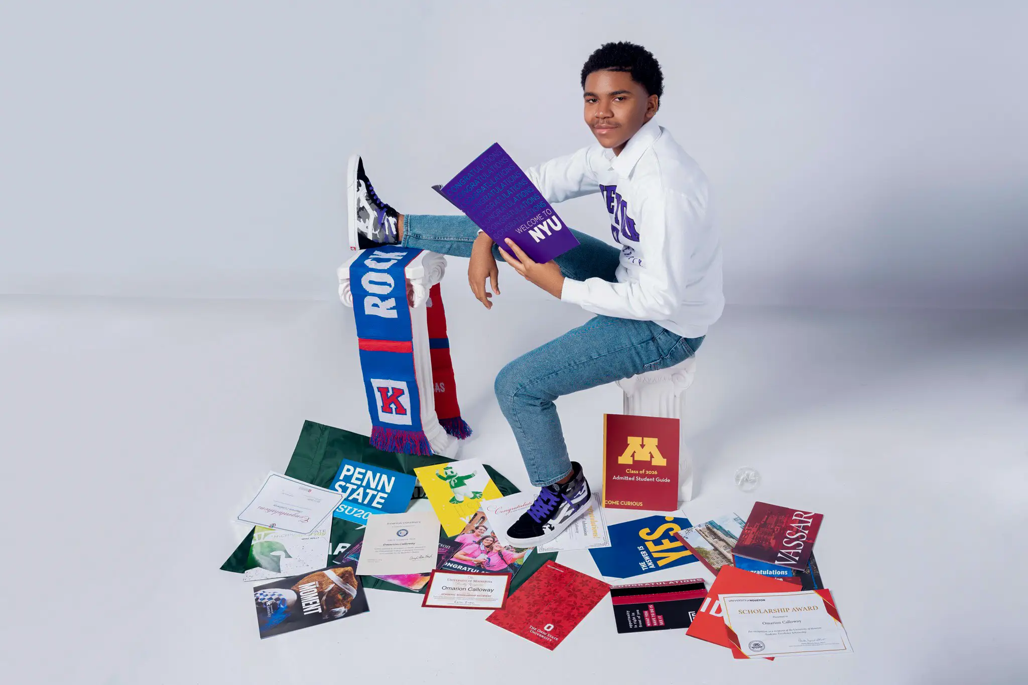 Student poses in a studio surrounded by college admissions booklets, certificates, and school-branded materials.
