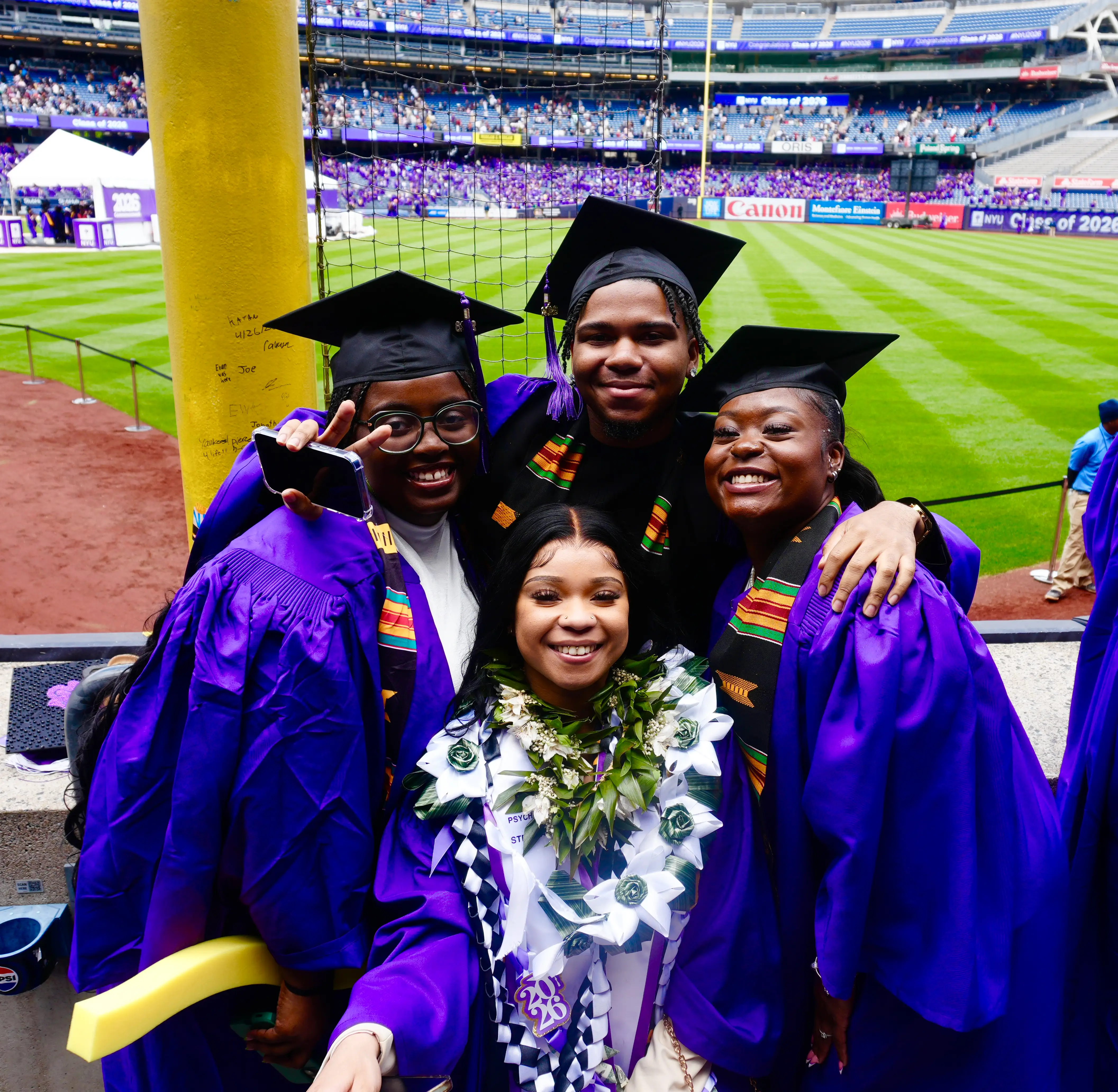 Four graduates in purple caps and gowns pose smiling beside a baseball field during an NYU Class of 2026 ceremony.