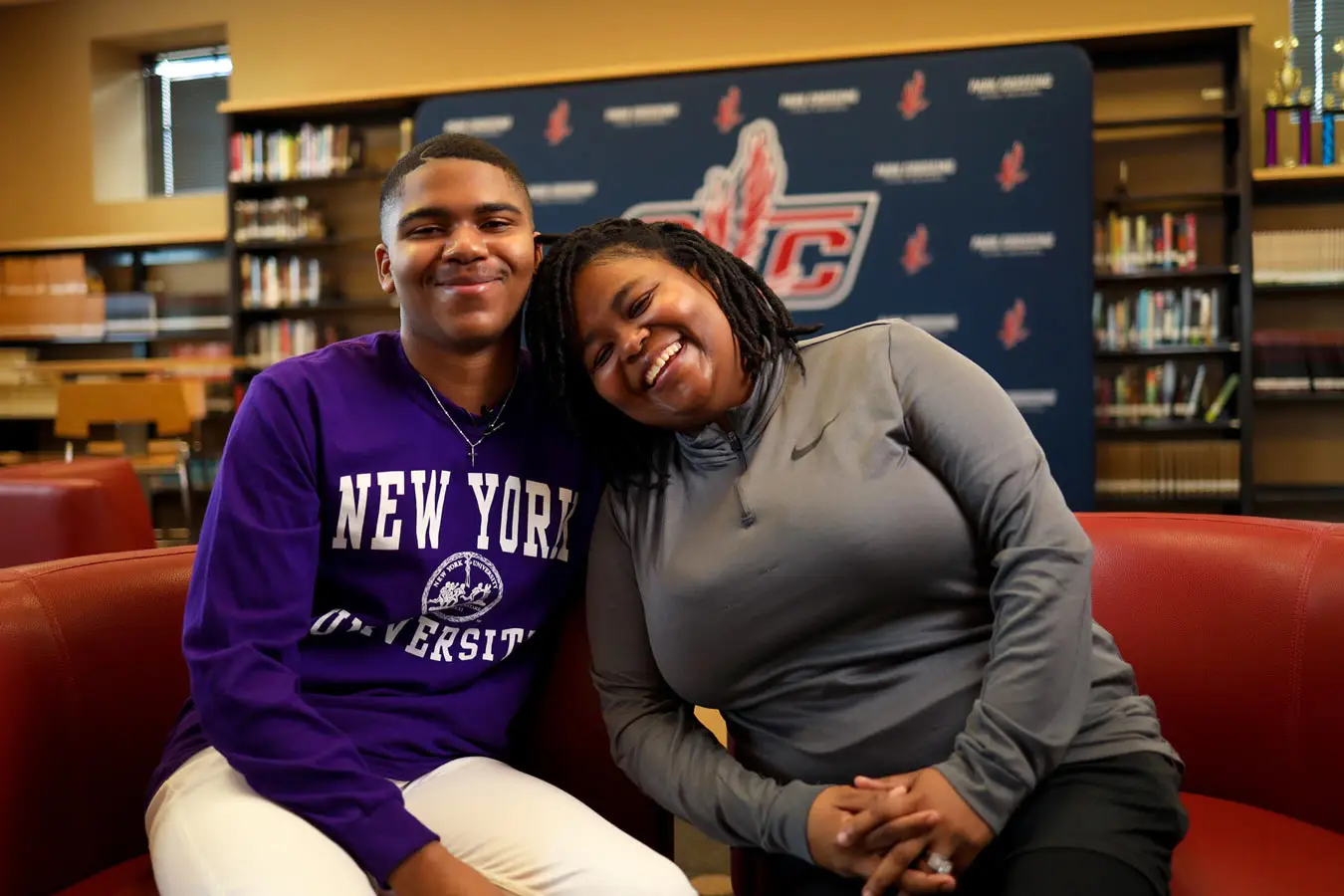 Two smiling people sit on a red couch in a library in front of a school-branded backdrop.