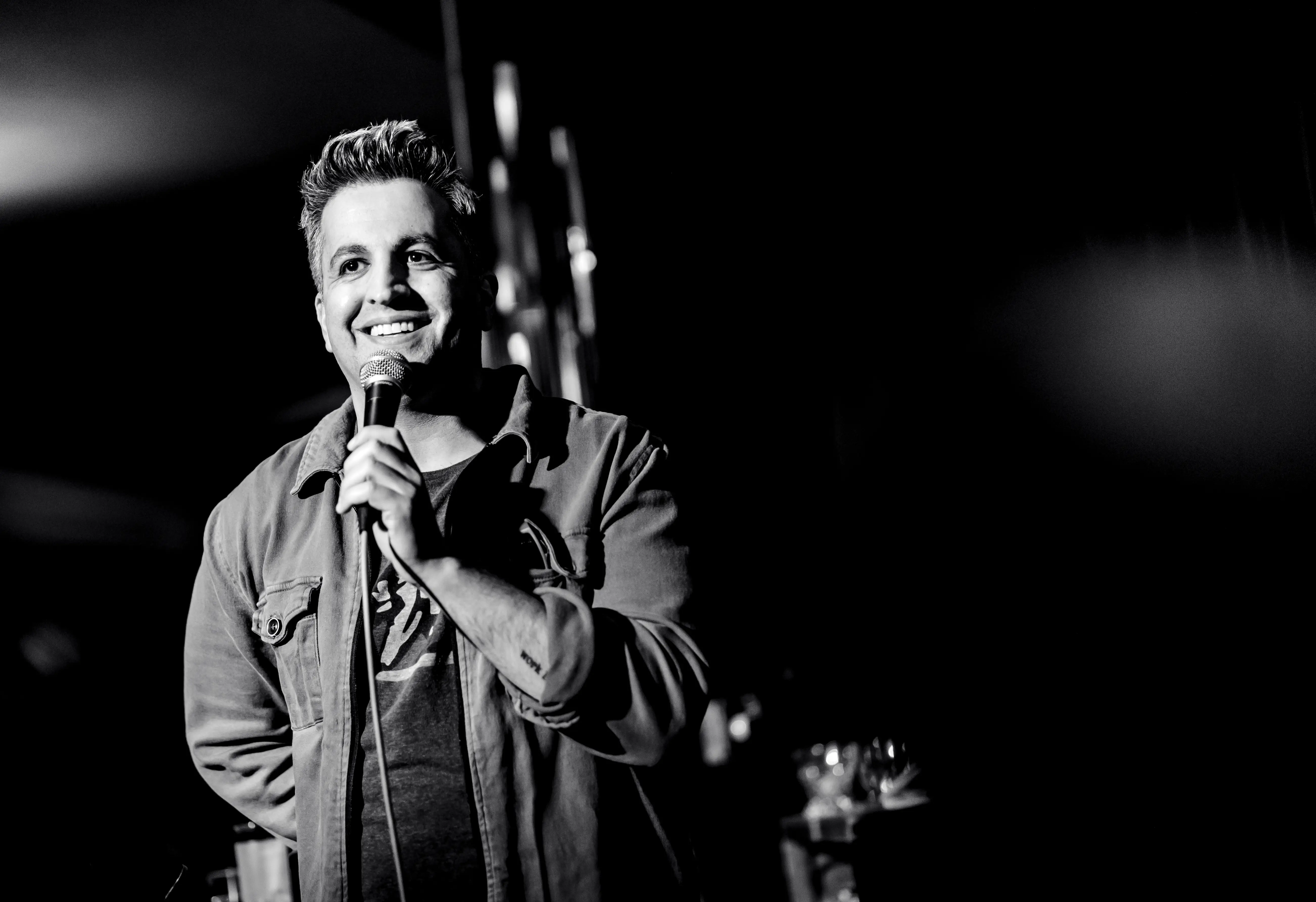 Paul Farahvar holds a microphone on a dark stage in a black-and-white live event photograph.