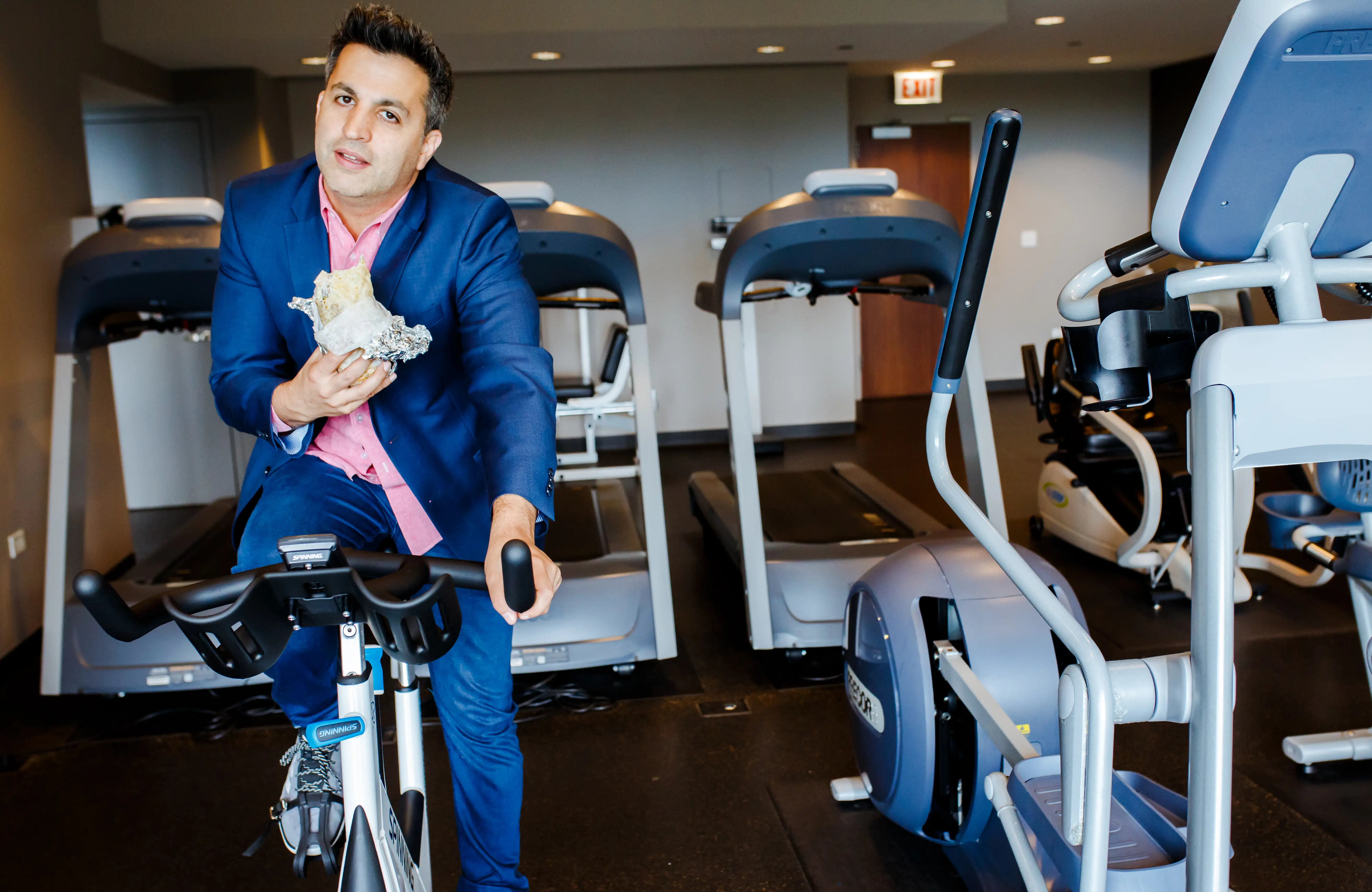 Paul Farahvar in a blue suit eats a wrapped sandwich while riding a stationary bike in a fitness room.