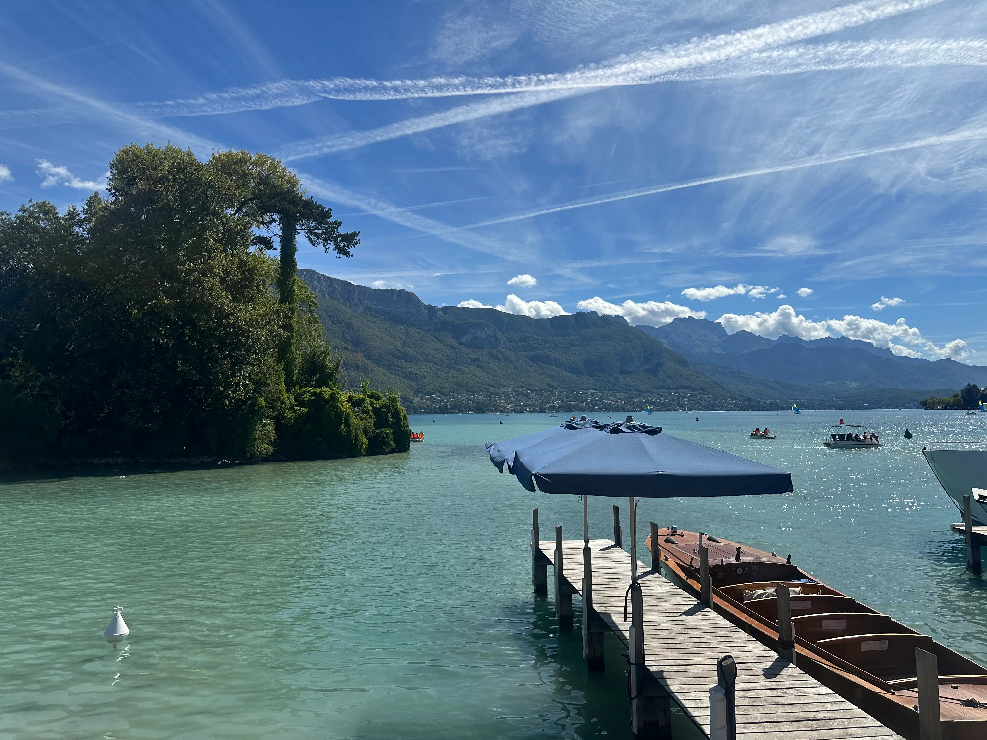 A wooden dock with a canopy extends into a turquoise lake with boats, mountains, trees, and streaked clouds.