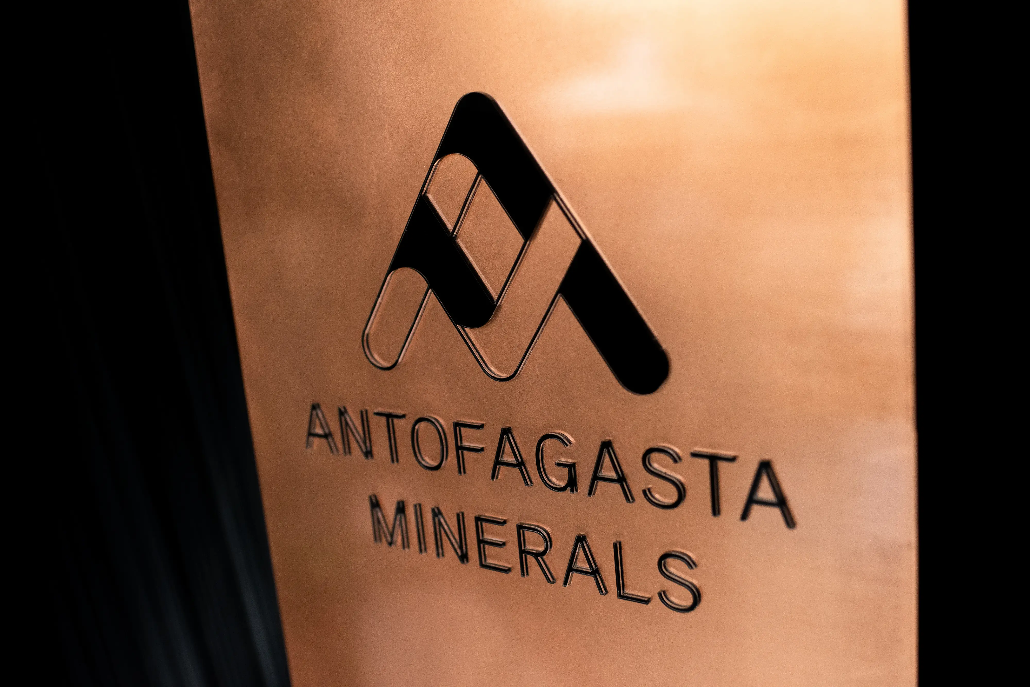 Copper-toned sign displays the Antofagasta Minerals logo and name against a dark background.
