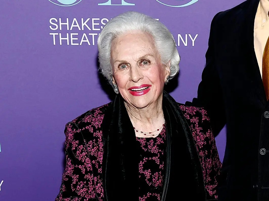 Two attendees pose on a purple Shakespeare Theatre Company backdrop, one holding a black handbag and cane.