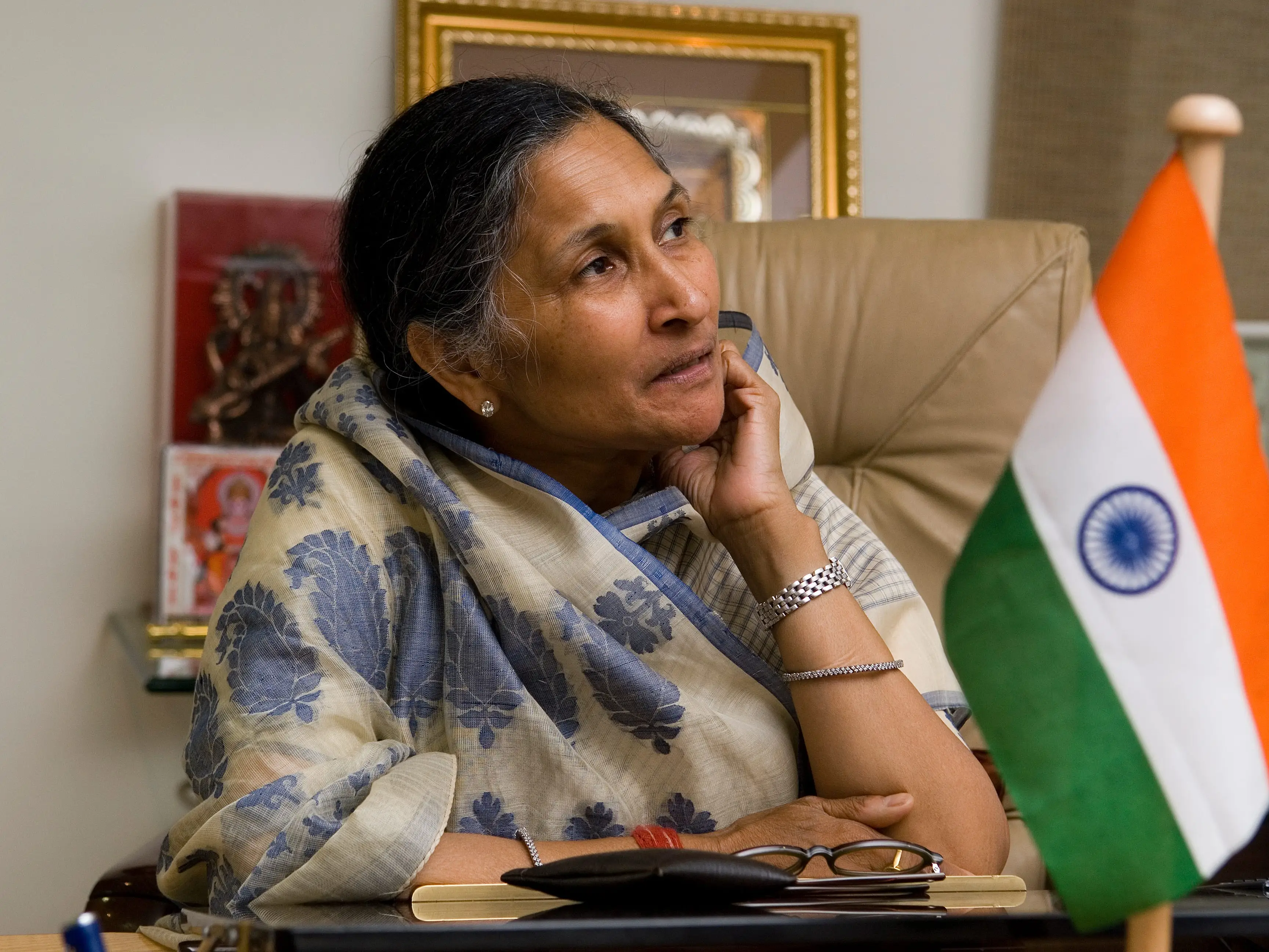 Person seated at an office desk beside an Indian national flag, with glasses and a notebook in the foreground.