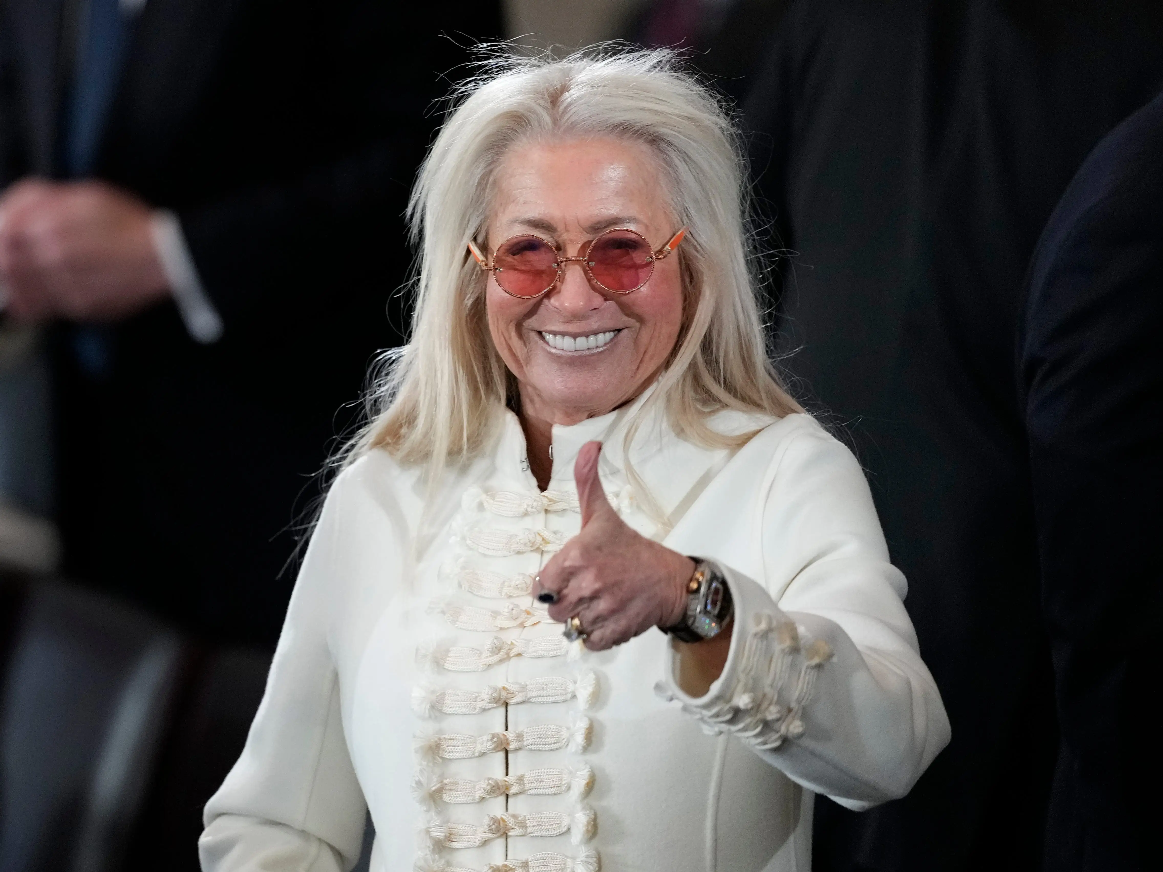 Miriam Adelson with long white hair, pink glasses, and a white jacket gives a thumbs-up among suited attendees.
