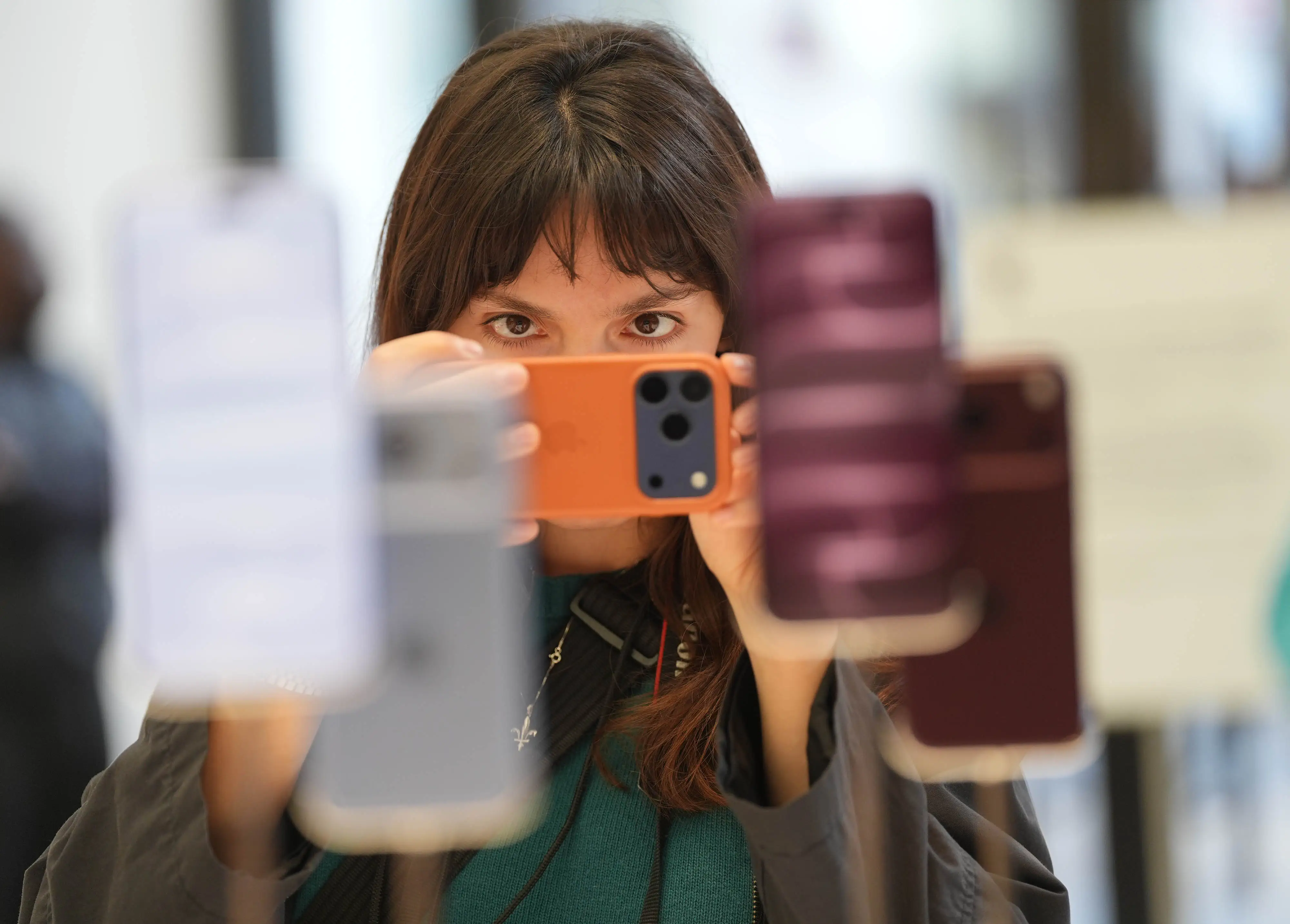 A shopper, holding an iPhone in an orange case, takes a horizontal picture of a display of iPhone 18 Pros.