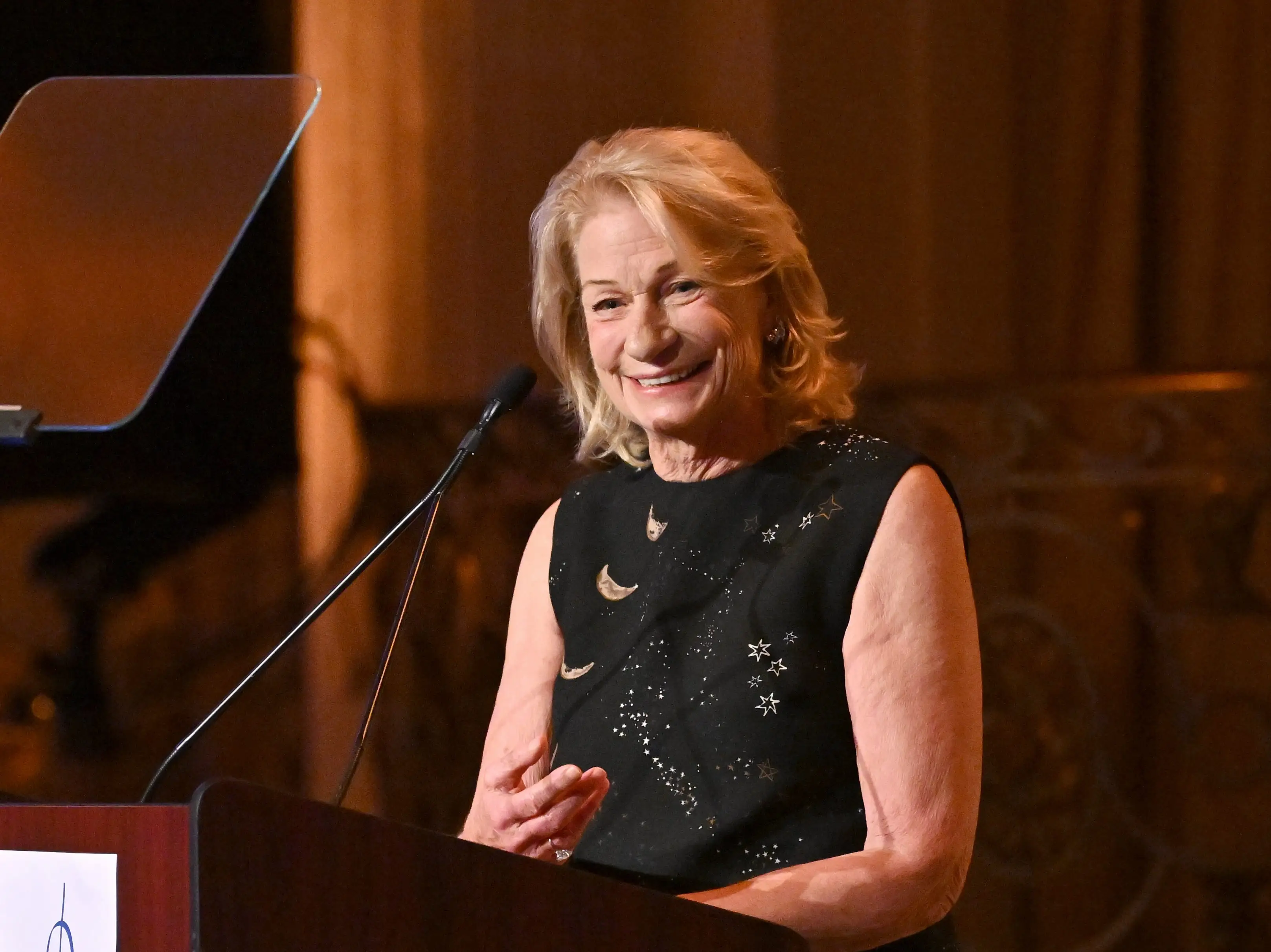 Marilyn Simons smiles while speaking at a podium with teleprompters in an ornate, warmly lit interior.