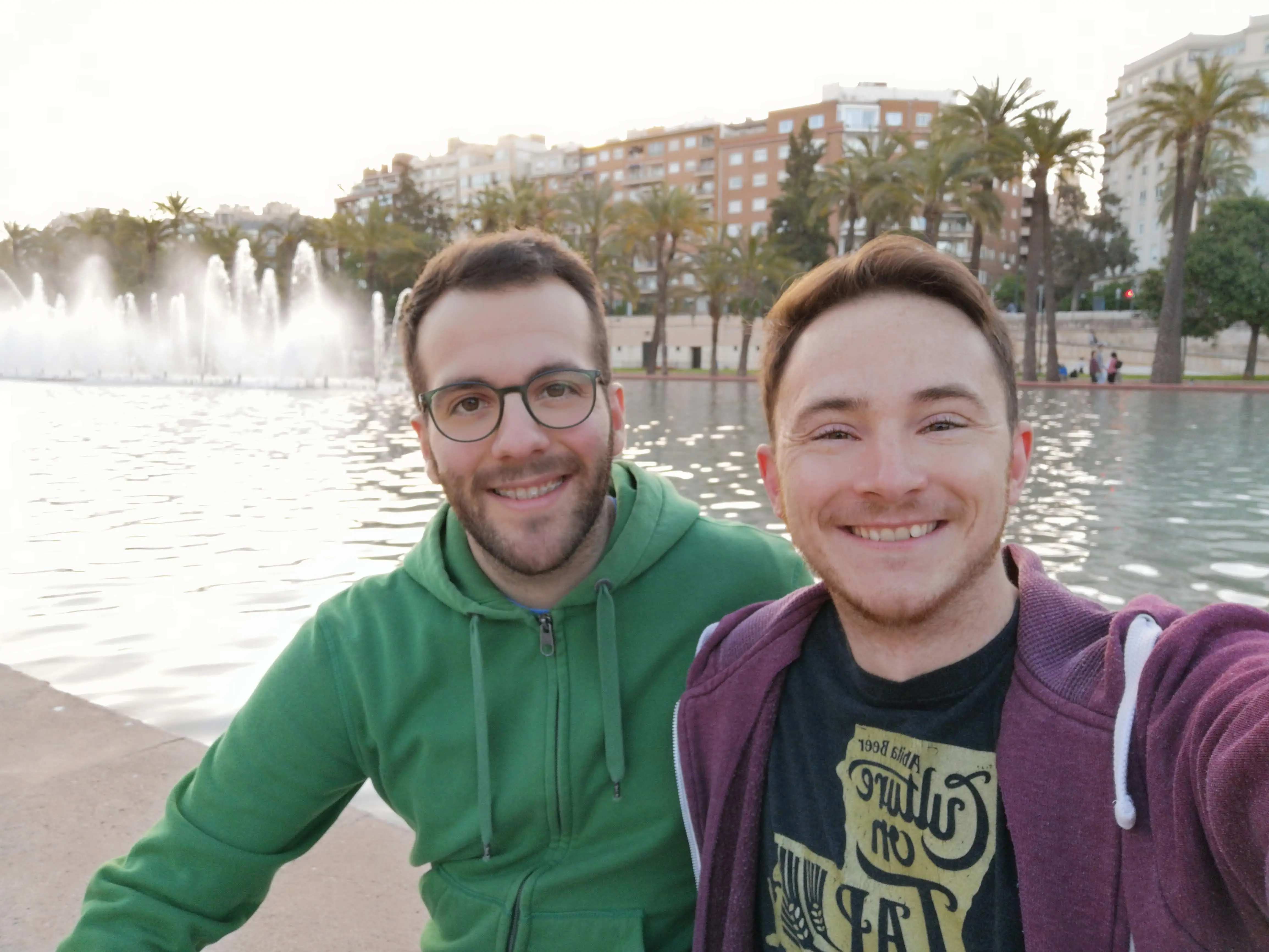 Two smiling people take a selfie beside a waterfront fountain with palm trees and city buildings behind them.