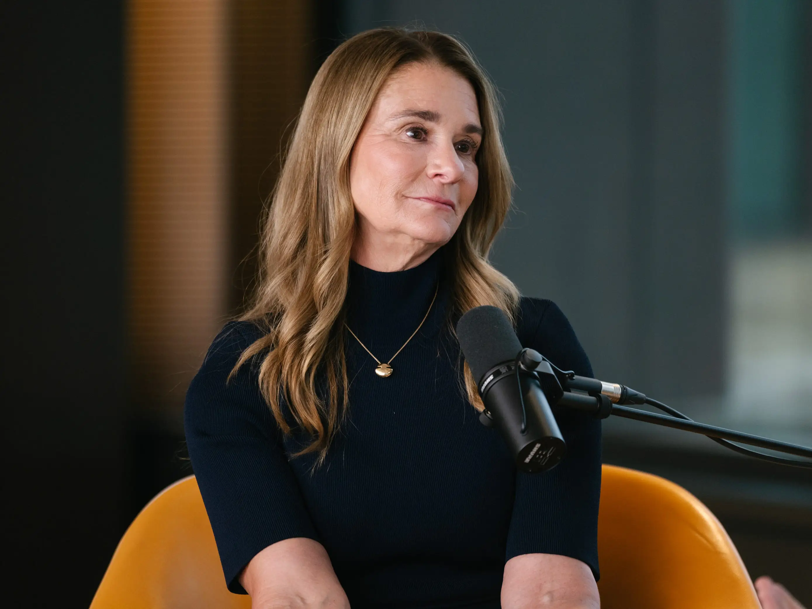 Melinda French Gates sits in an orange chair beside a studio microphone during an interview or podcast recording.