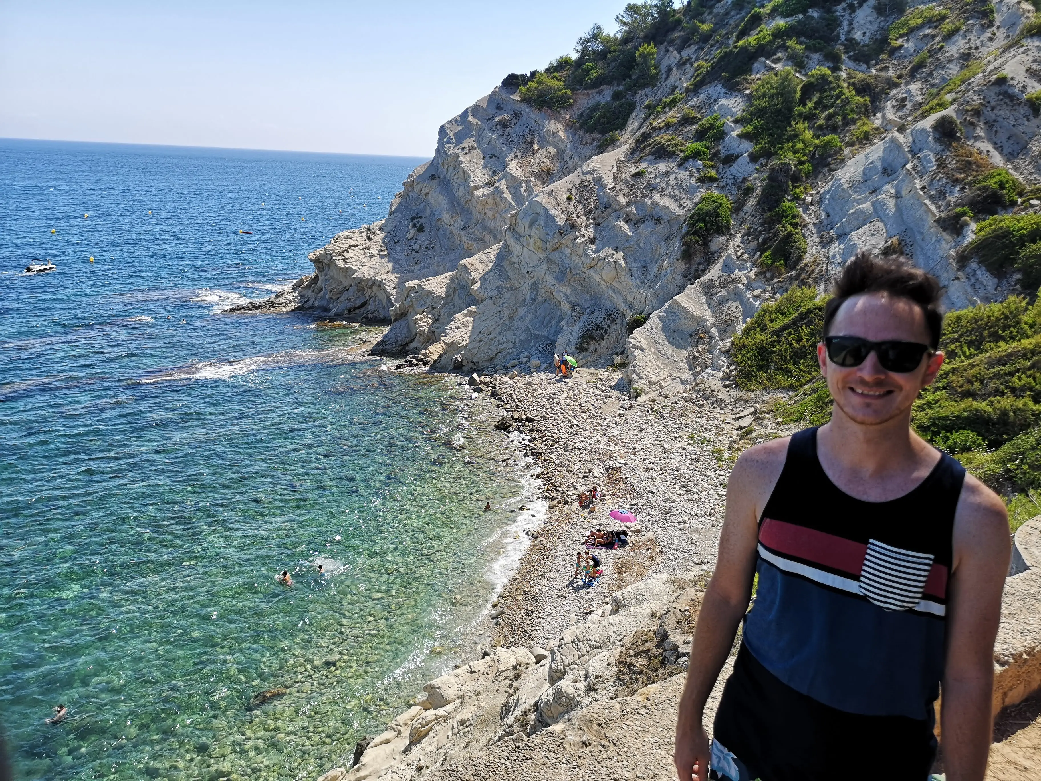 Kevin Joseph, wearing sunglasses, stands above a rocky beach with clear blue waters and white coastal cliffs.