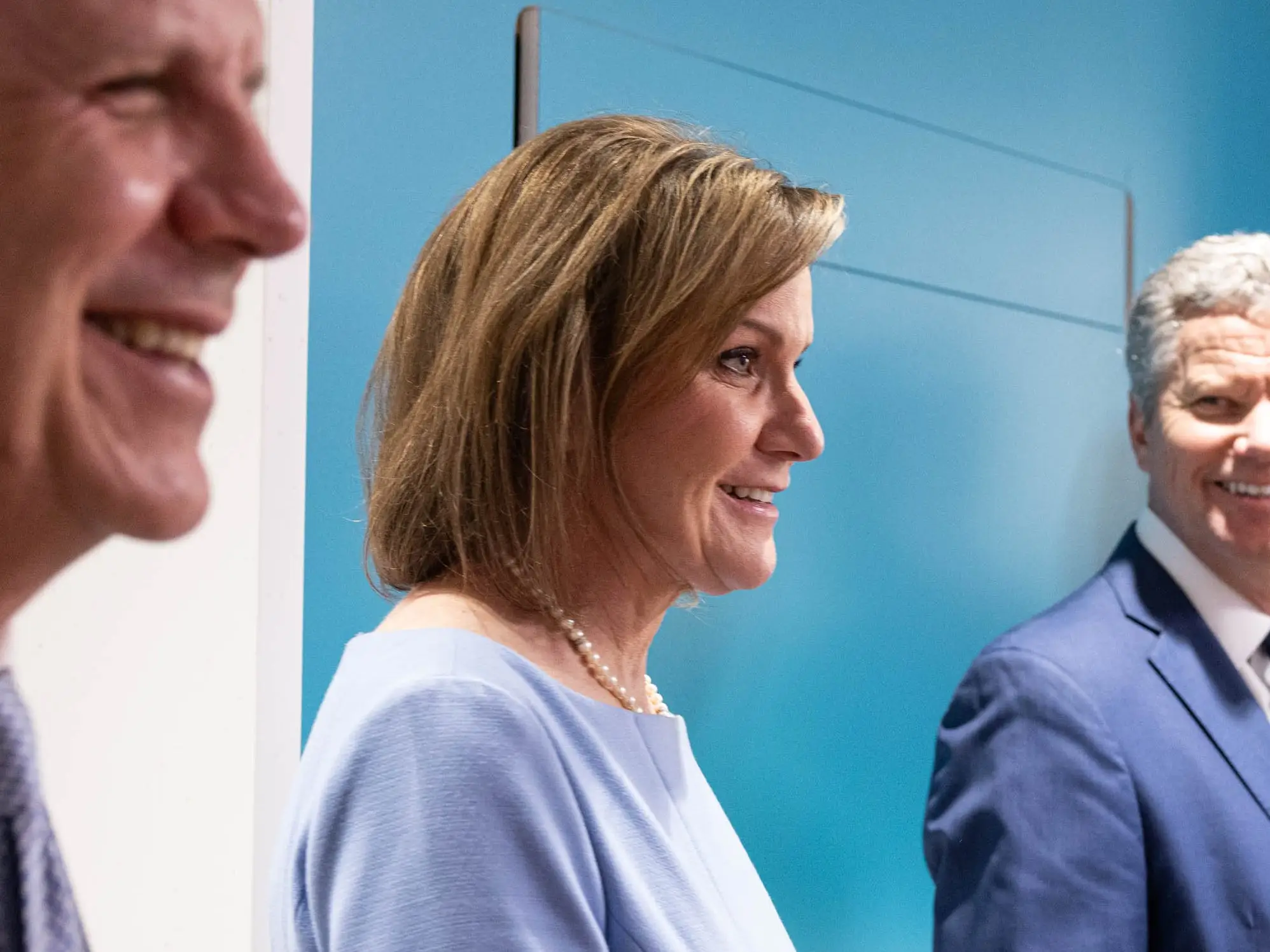 Five formally dressed people stand smiling in a bright hallway beside a teal wall.