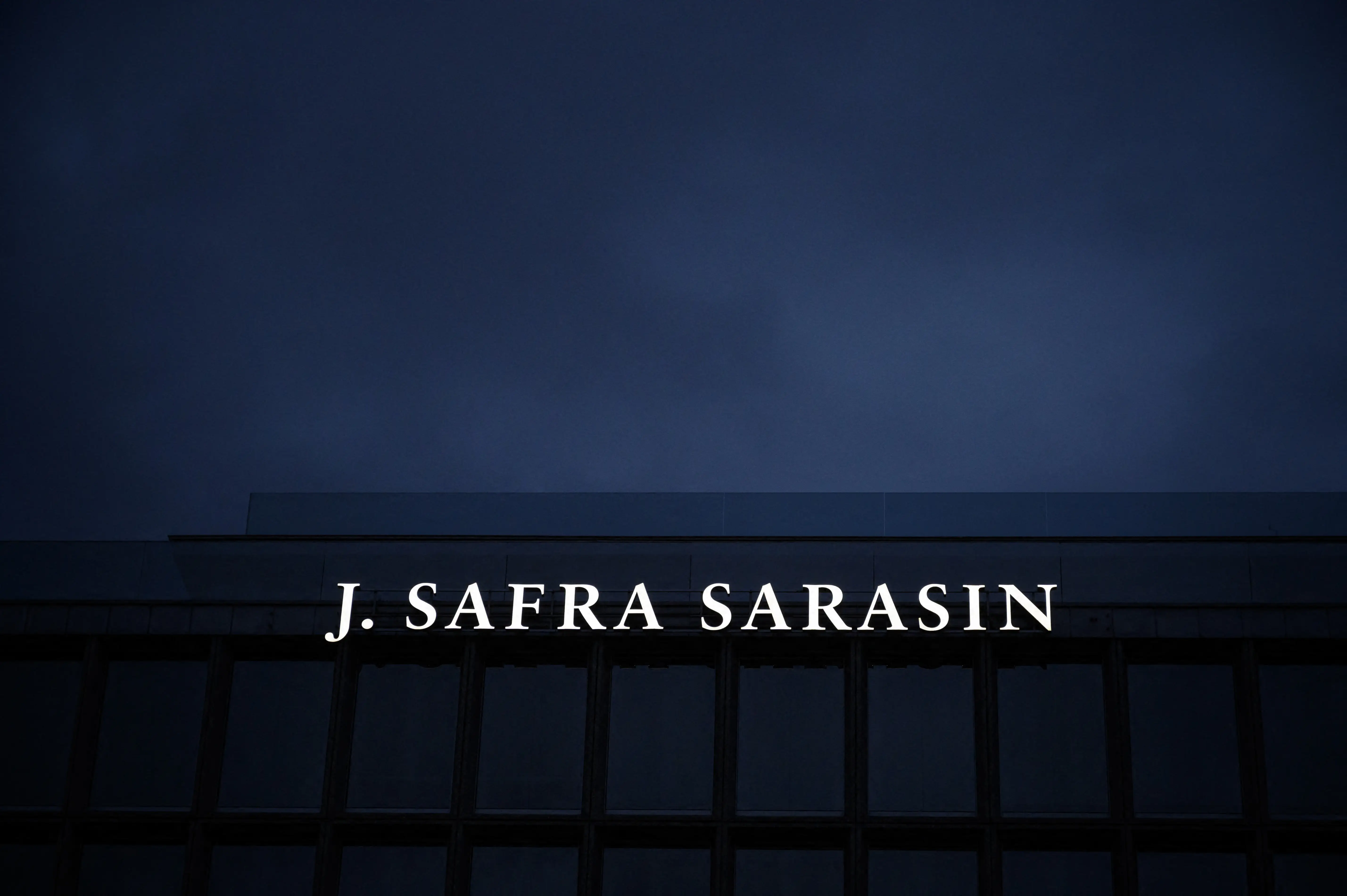 Illuminated J. SAFRA SARASIN sign on a dark building facade against a blue evening sky.