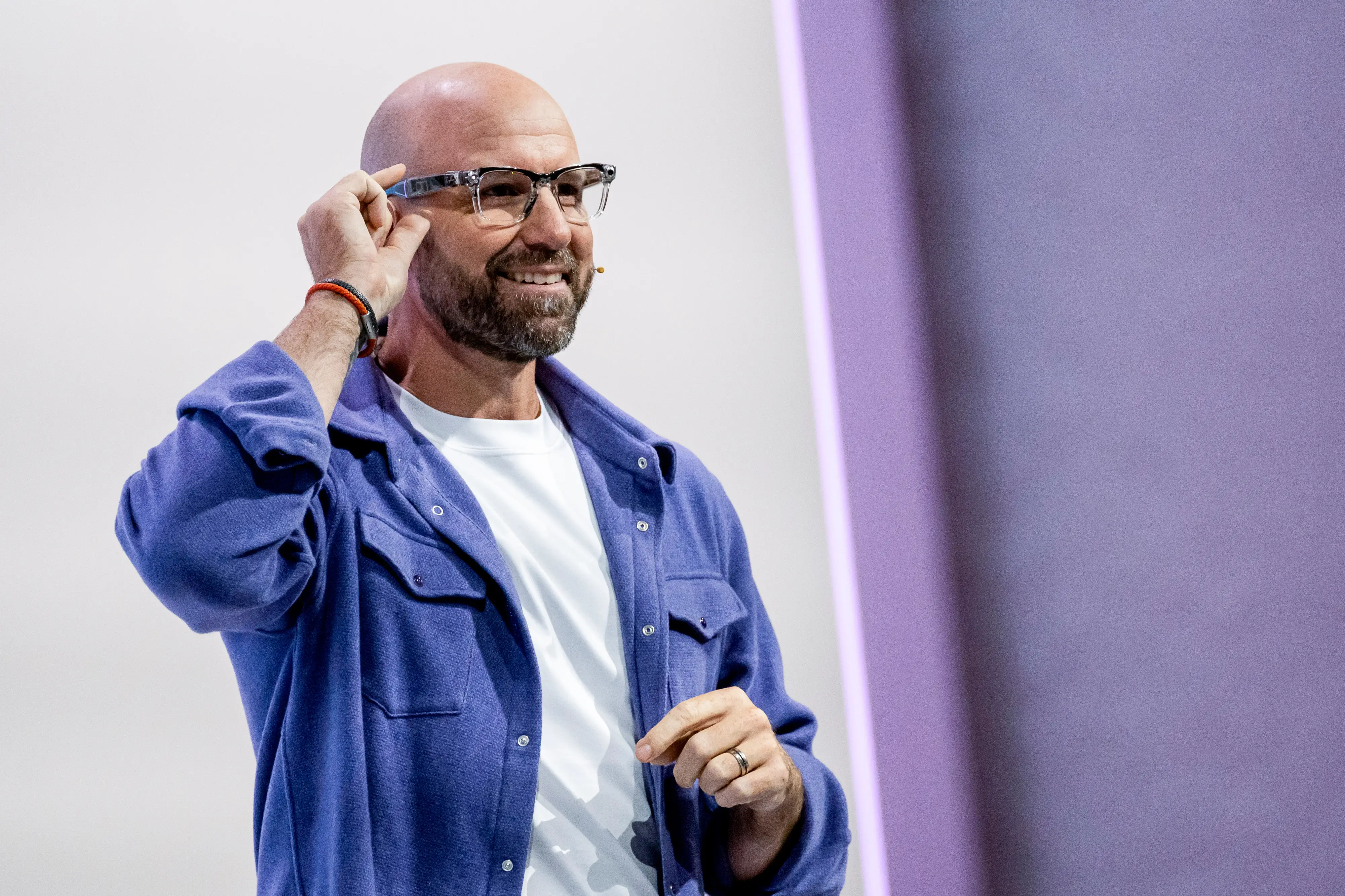 Smiling speaker in glasses and a blue overshirt adjusts eyewear on a brightly lit stage, andrew bosworth